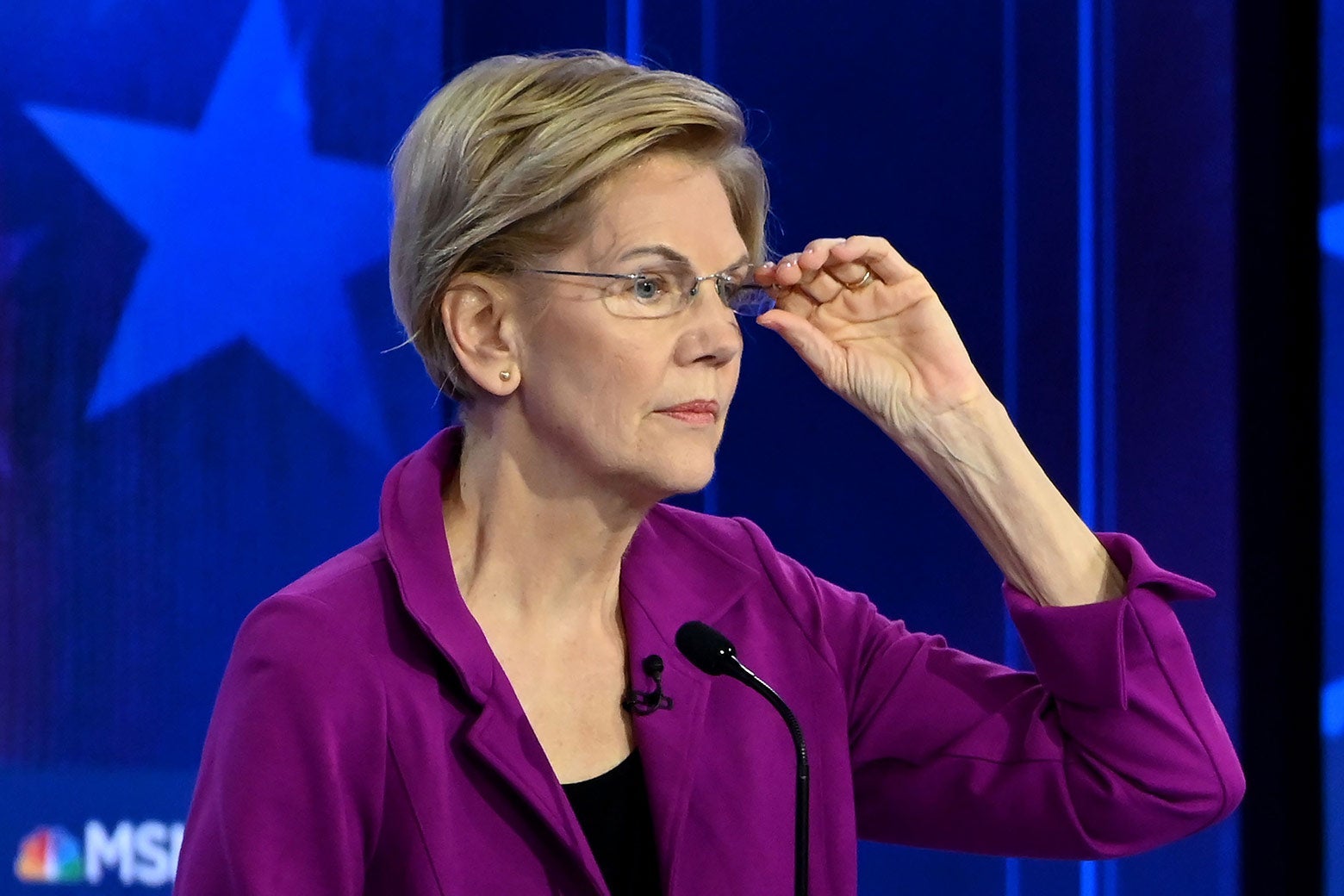 Elizabeth Warren adjusts her glasses while standing at a debate podium.