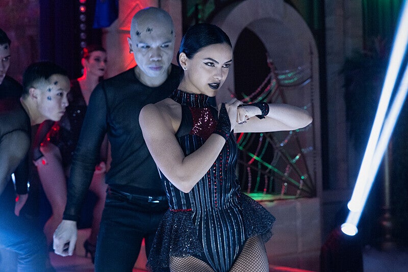 Camila Mendes holds a pose in a decorative black unitard while men in black costumes stand behind her. 