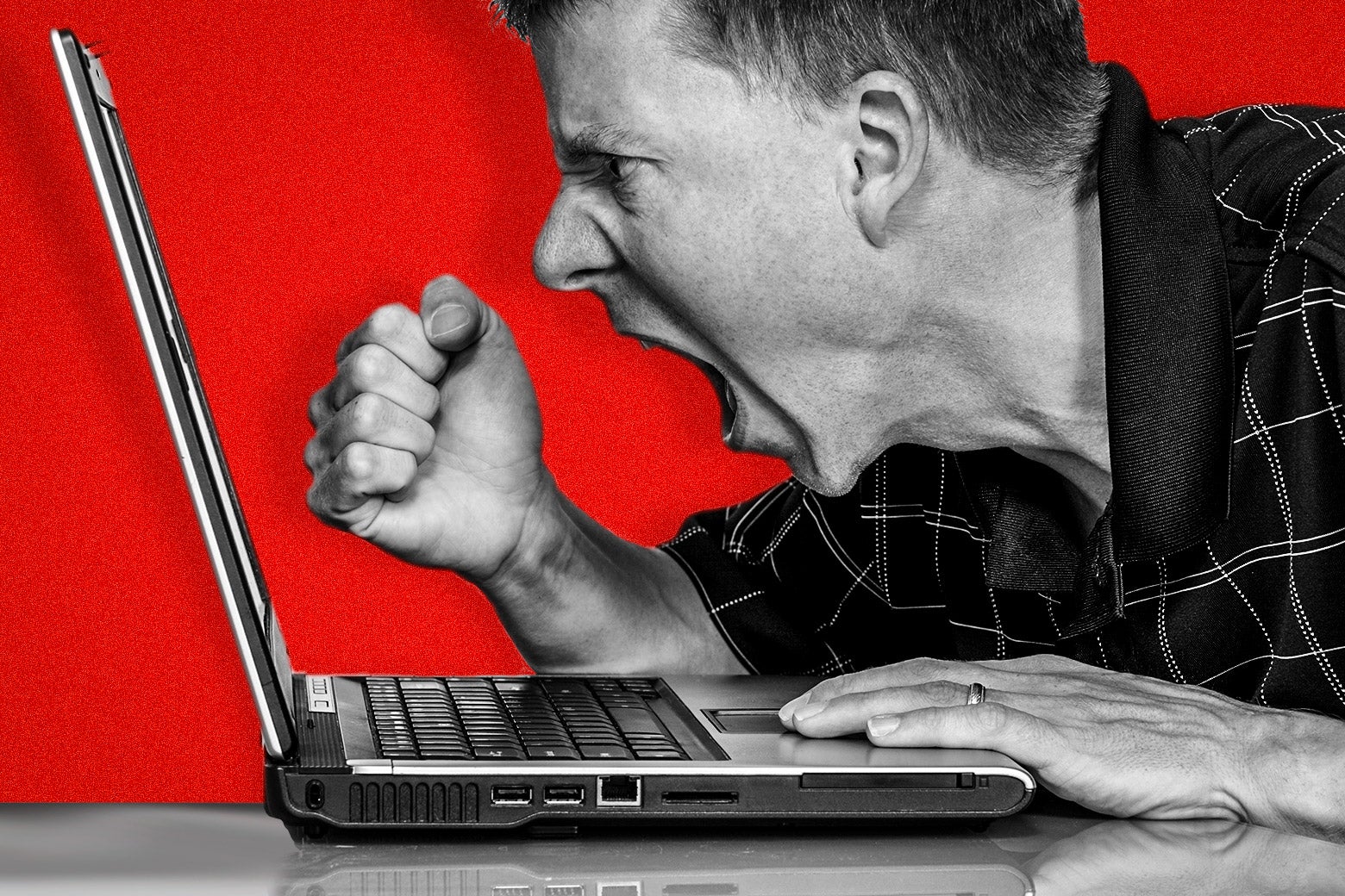 A man wearing a black checkered polo shirt shakes his fist and yells at his laptop screen.