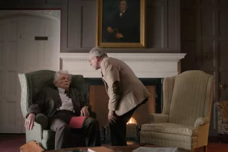 Elliot talks to a sleeping colleague sitting in an armchair.