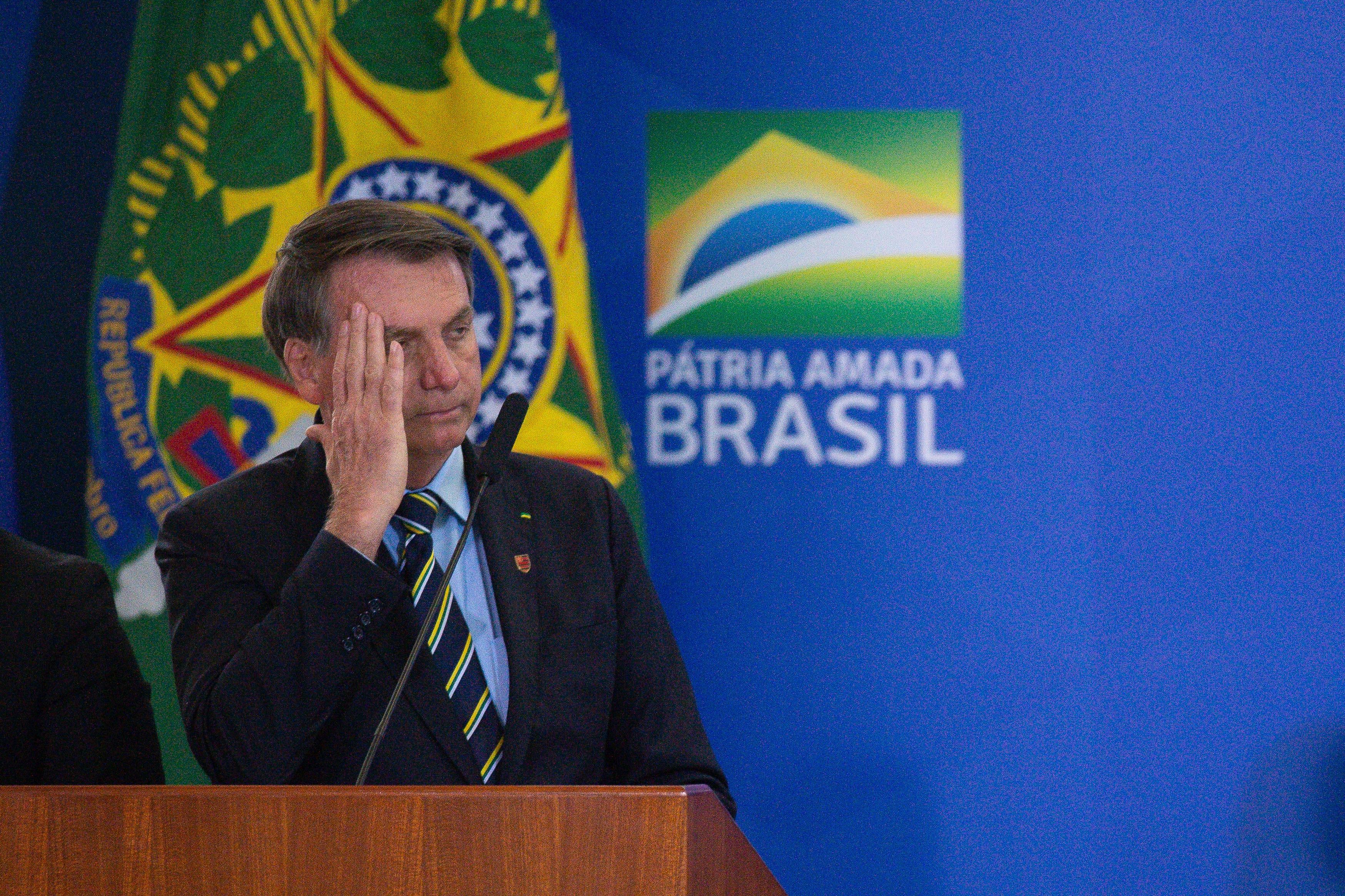 Bolsonaro, at a lectern, holds a hand by his head.