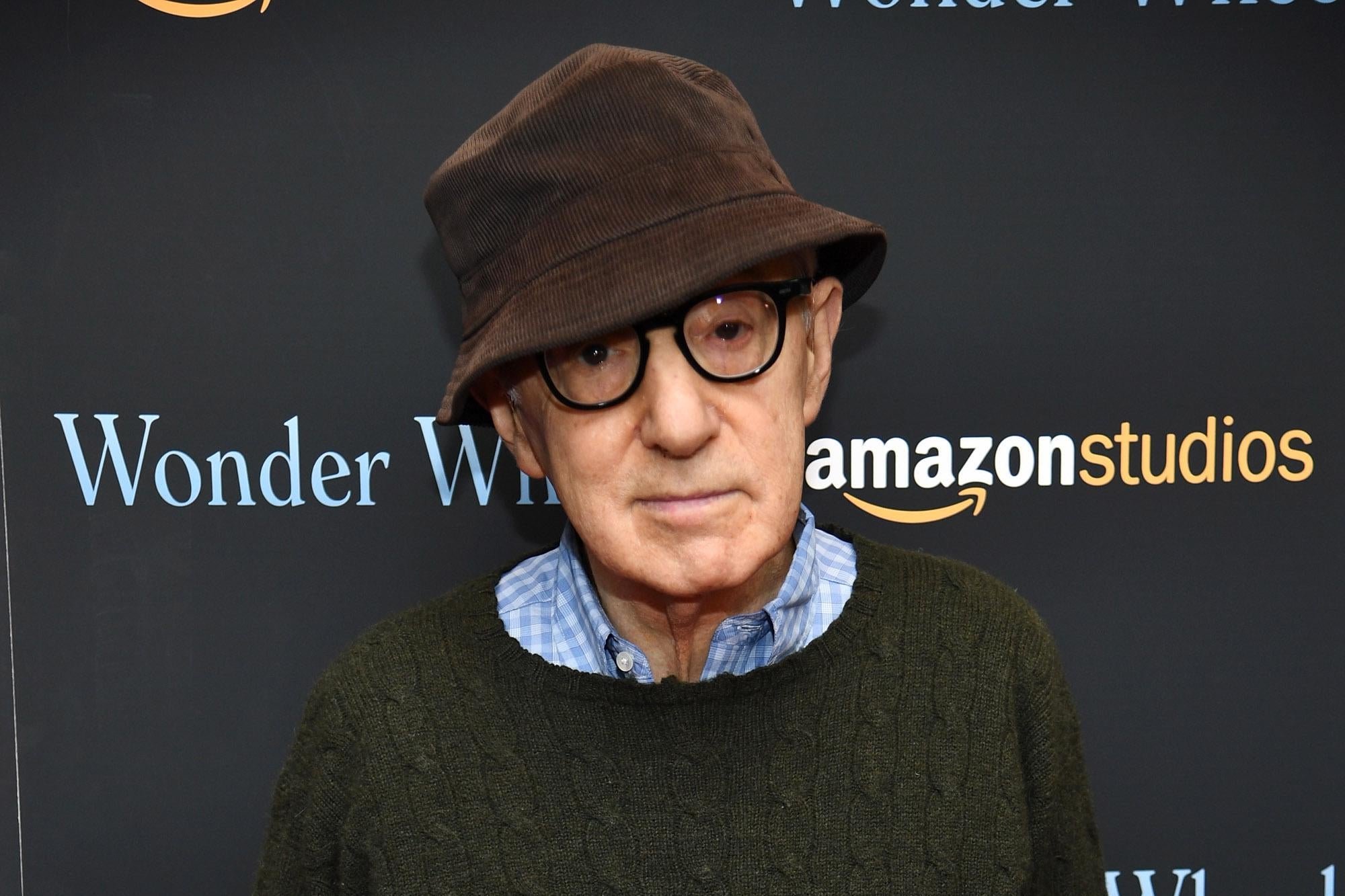 Woody Allen wears a brown bucket hat and black glasses as he poses in front of a backdrop the says "Wonder Wheel" and "Amazon Studios."
