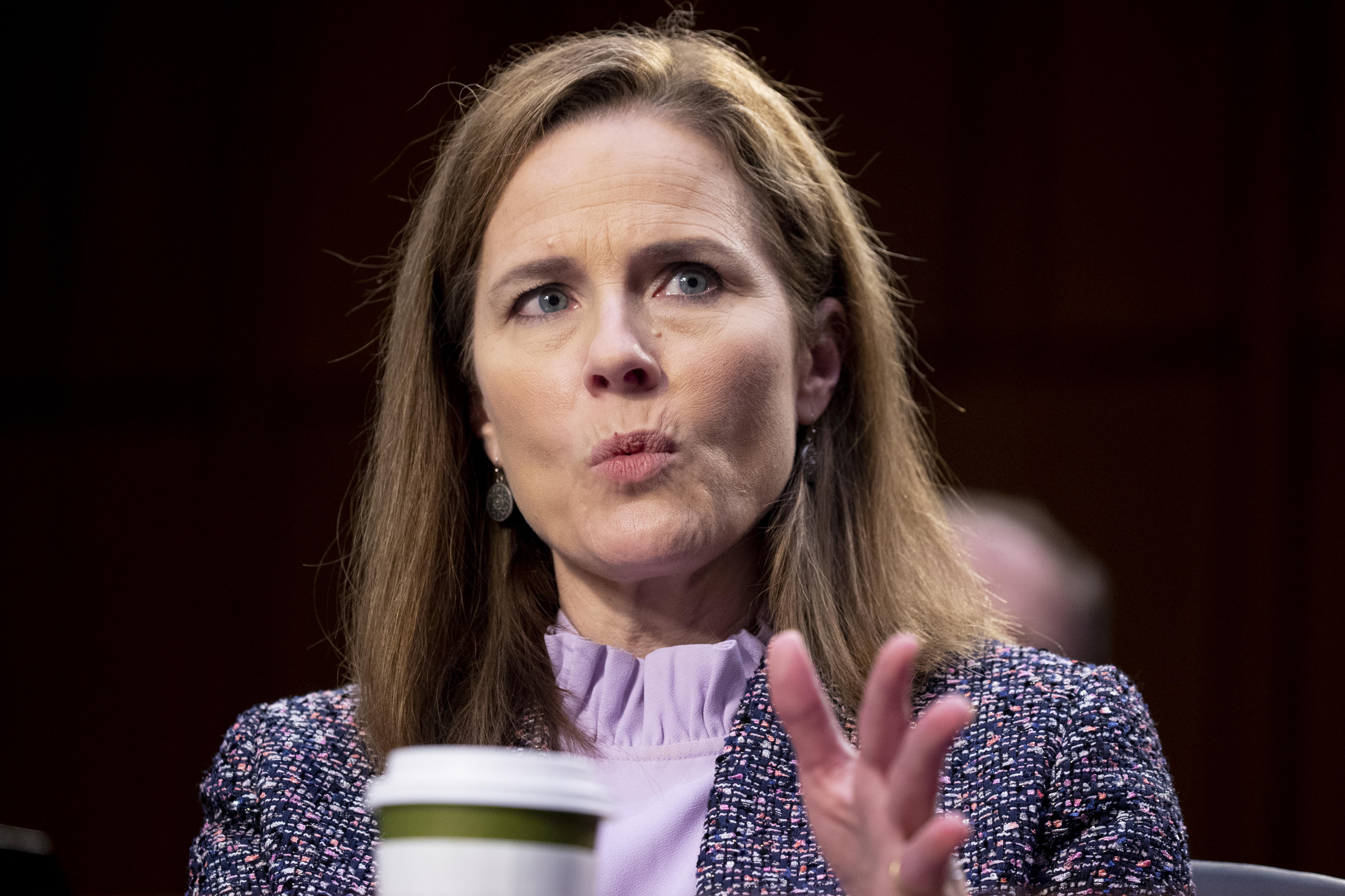 Amy Coney Barrett speaks while gesturing. A coffee cup can be seen in front of her.