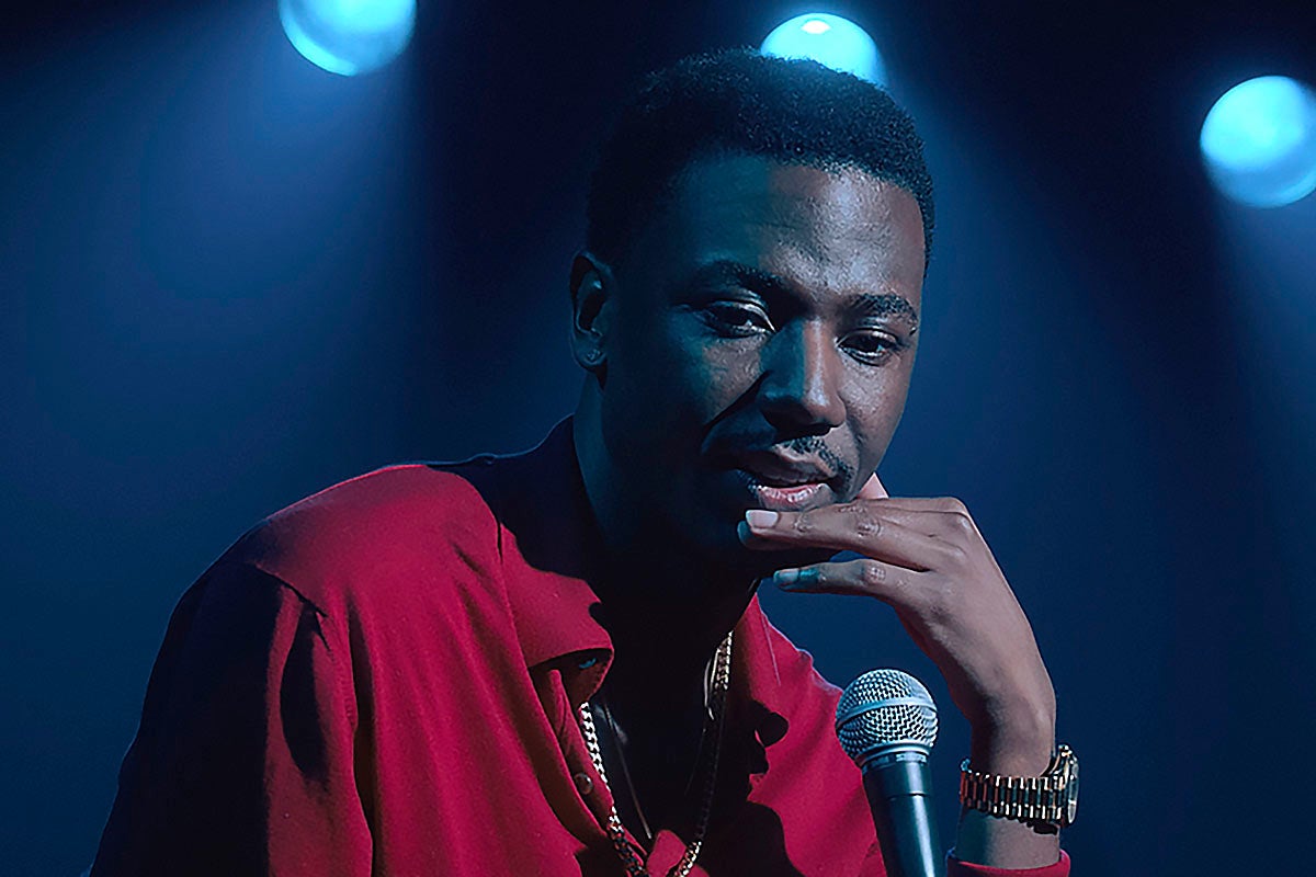 Jerrod Carmichael sits at the mic in a red shirt looking pensive.