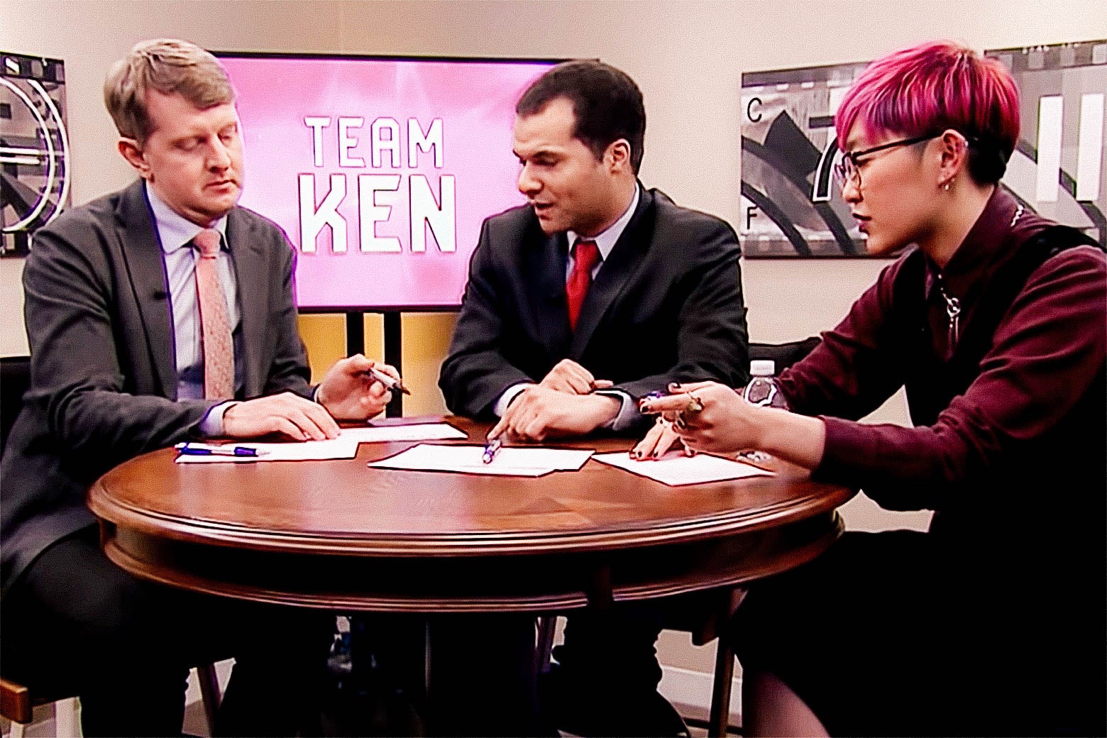 Ken Jennings, Matt Jackson, and Monica Thieu conferring at a round table. A TV screen in the background reads "TEAM KEN."