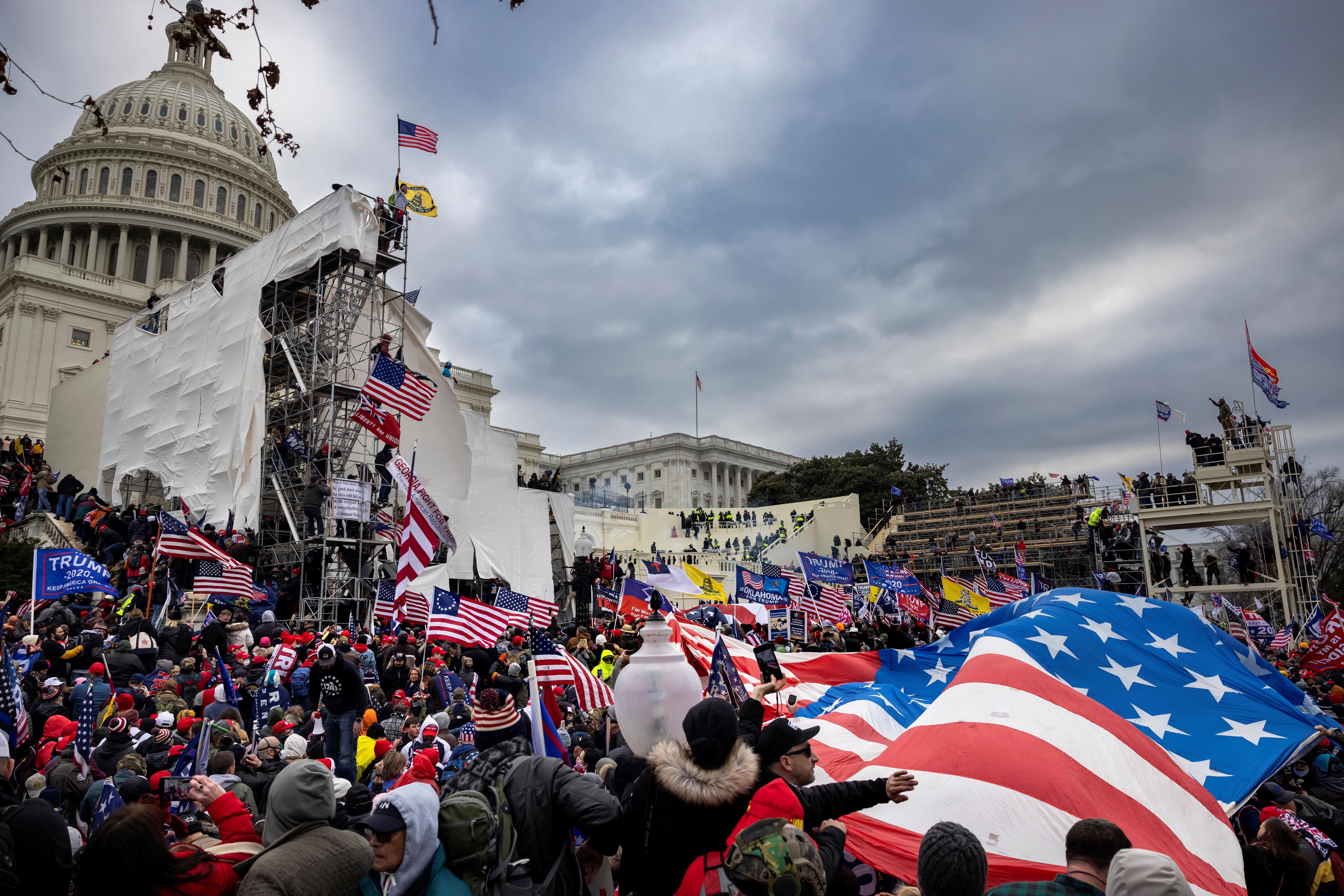 A huge American flag billows in the lower right foreground, overlooked by the Capitol dome in the upper left background, as a crowd bearing various U.S. and Trump flags swarms around the inauguration scaffolding outside the Capitol.