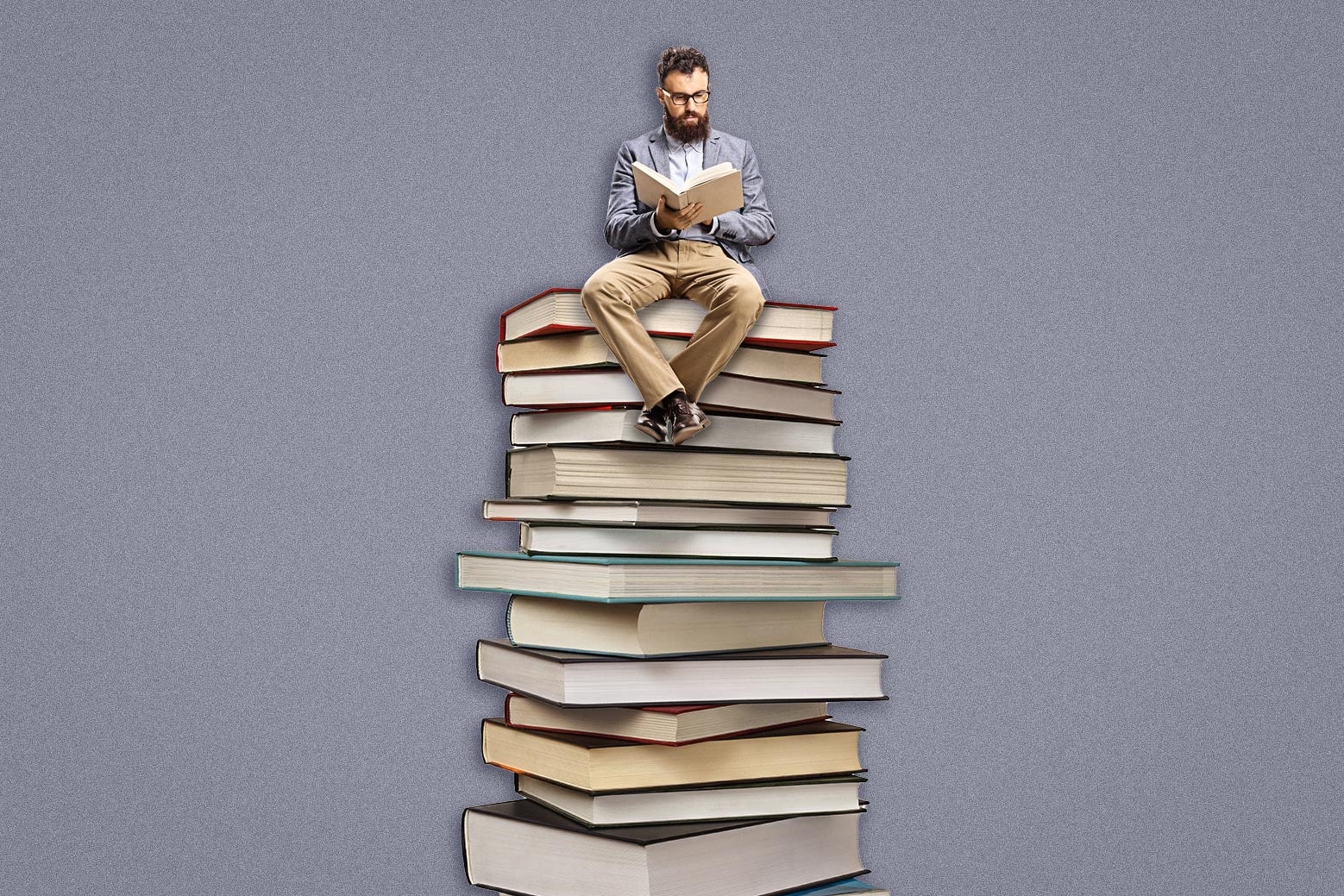 A man in a professor-ish sportscoat sits reading on top of a stack of books.