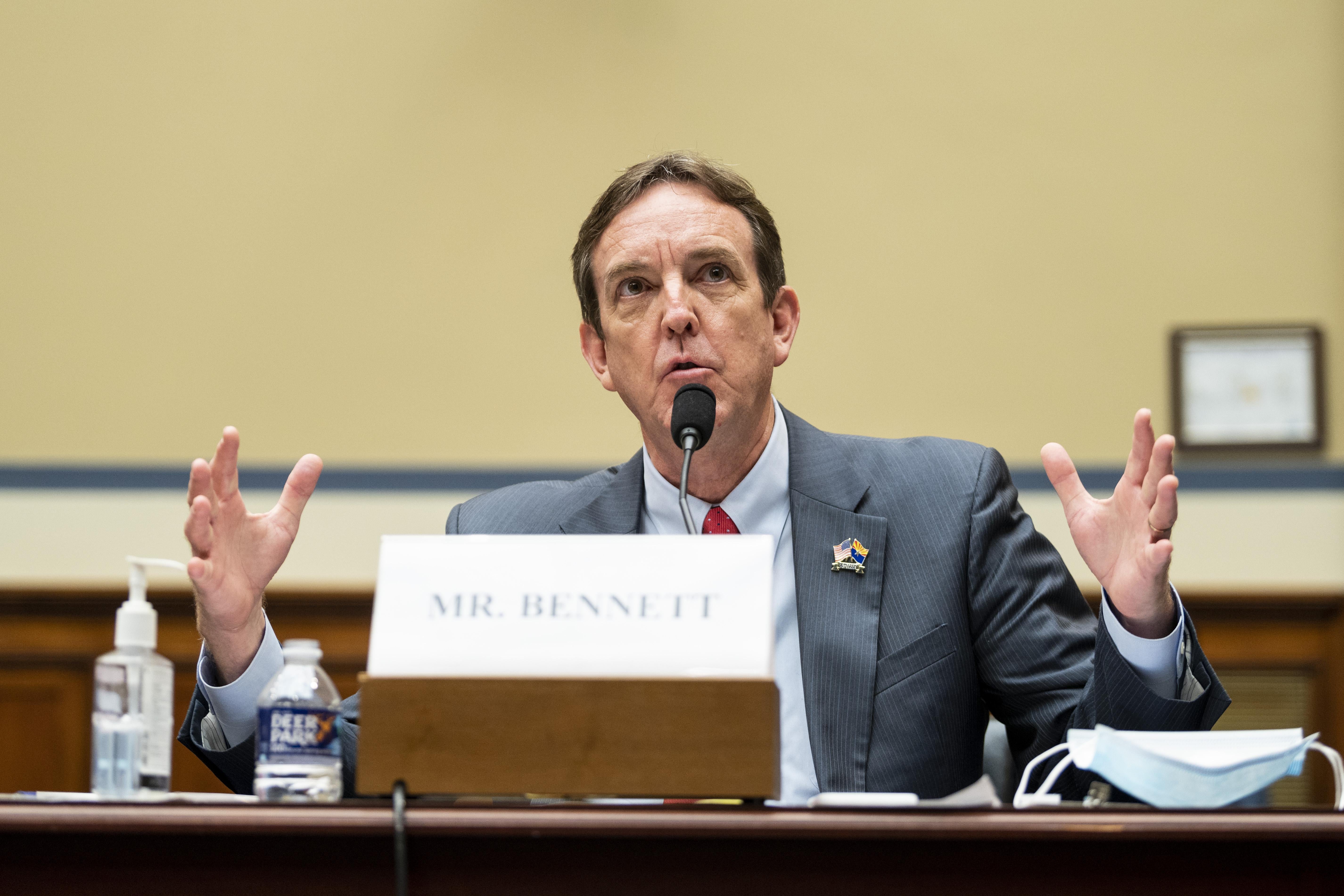Ken Bennett gestures with both hands as he speaks at a mic while seated on a panel
