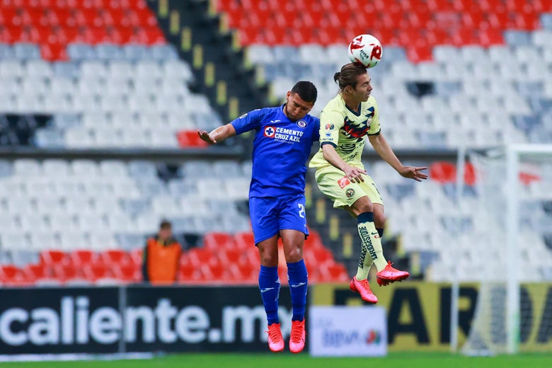 A soccer player heads a ball as another player jumps behind him, in an empty stadium.