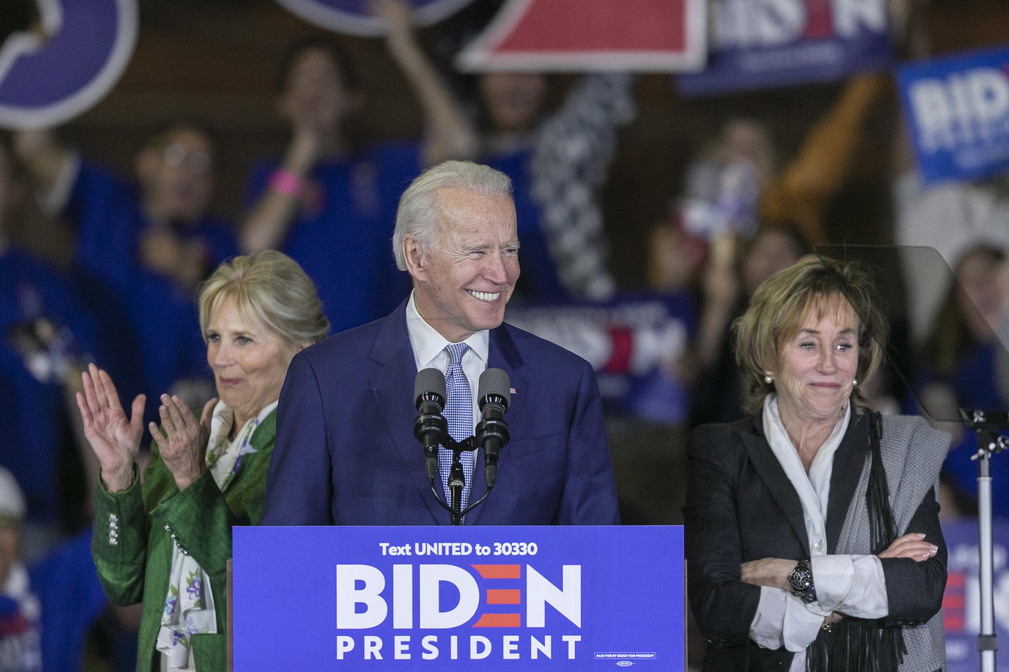 Joe Biden is smiling at a rally behind a podium that says BIDEN PRESIDENT, standing next to his wife Jill Biden and sister Valerie Biden Owens. People with Biden signs cheer in the background.