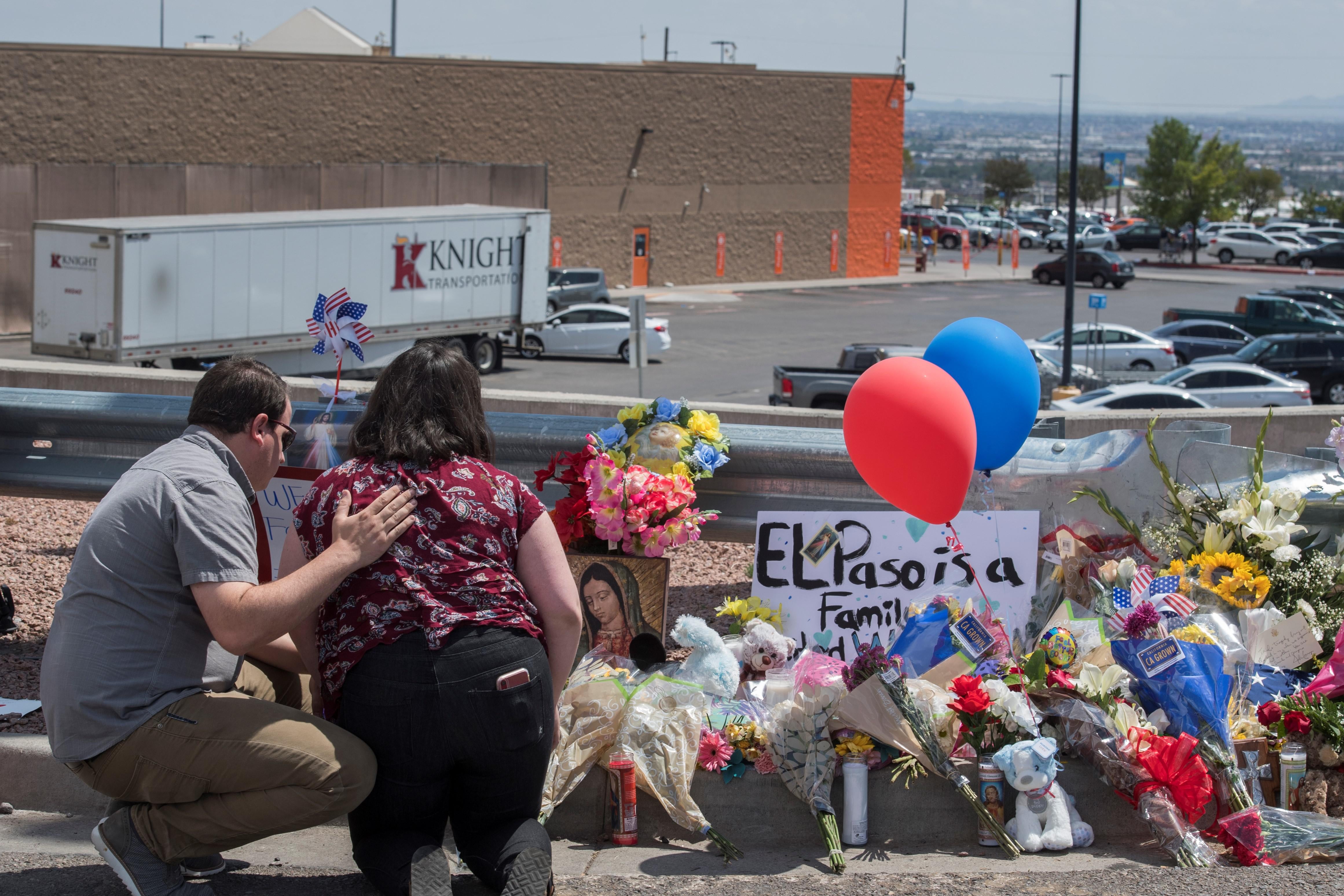 Two people kneel beside a memorial of flowers, balloons, and religious items honoring the victims of the shooting. A sign says "El Paso is a family."