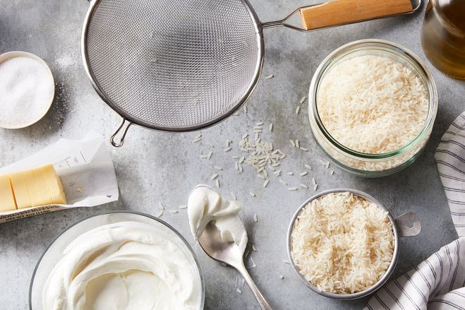 On a countertop, jars of dry rice, a sieve, yogurt, salt, butter, and a spoon.
