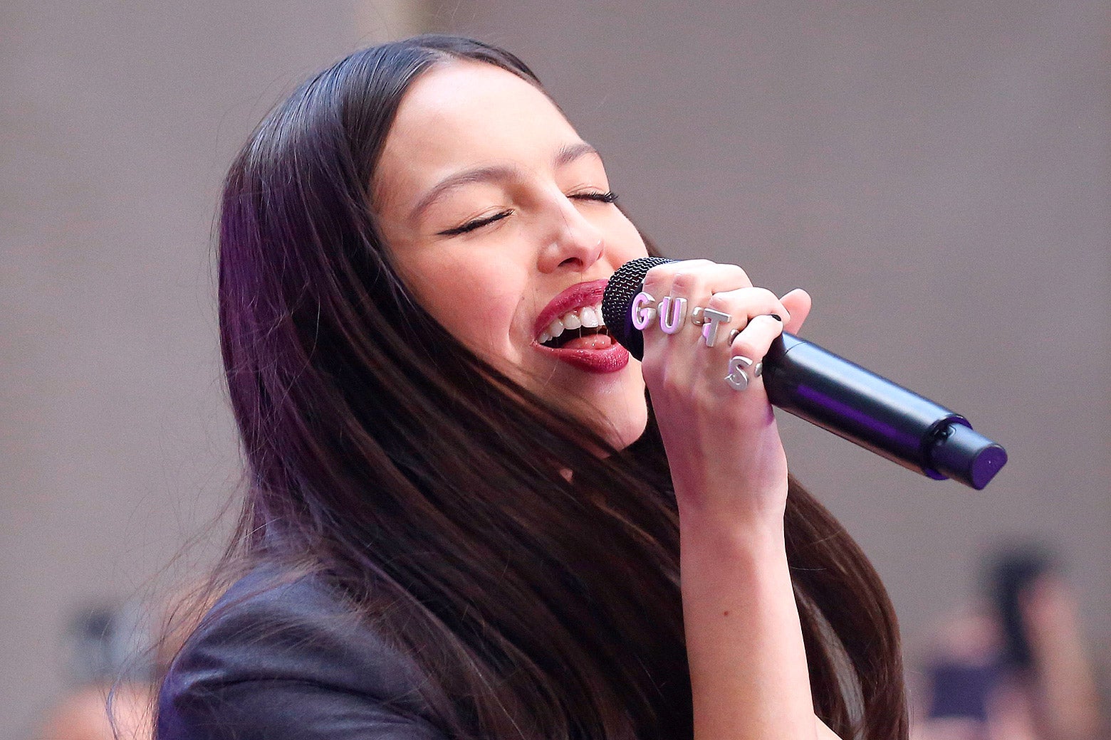 The young singer smiles as she closes her eyes and lifts a microphone to her lips, wearing red lipstick and her long dark hair in a center part.