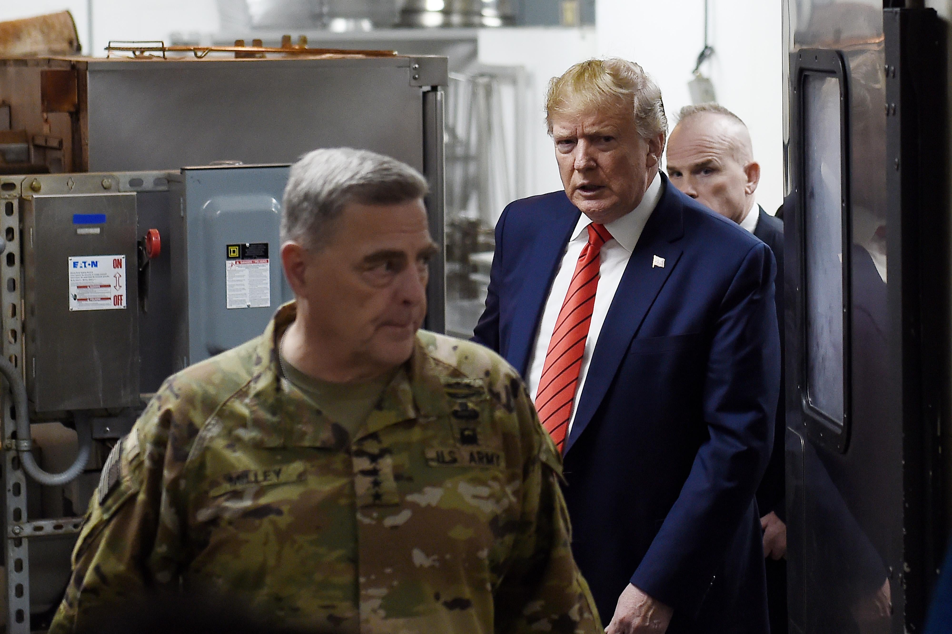 Mark Milley, wearing camo, stands in front of Donald Trump, who's in a suit. Another man can be seen behind Trump.