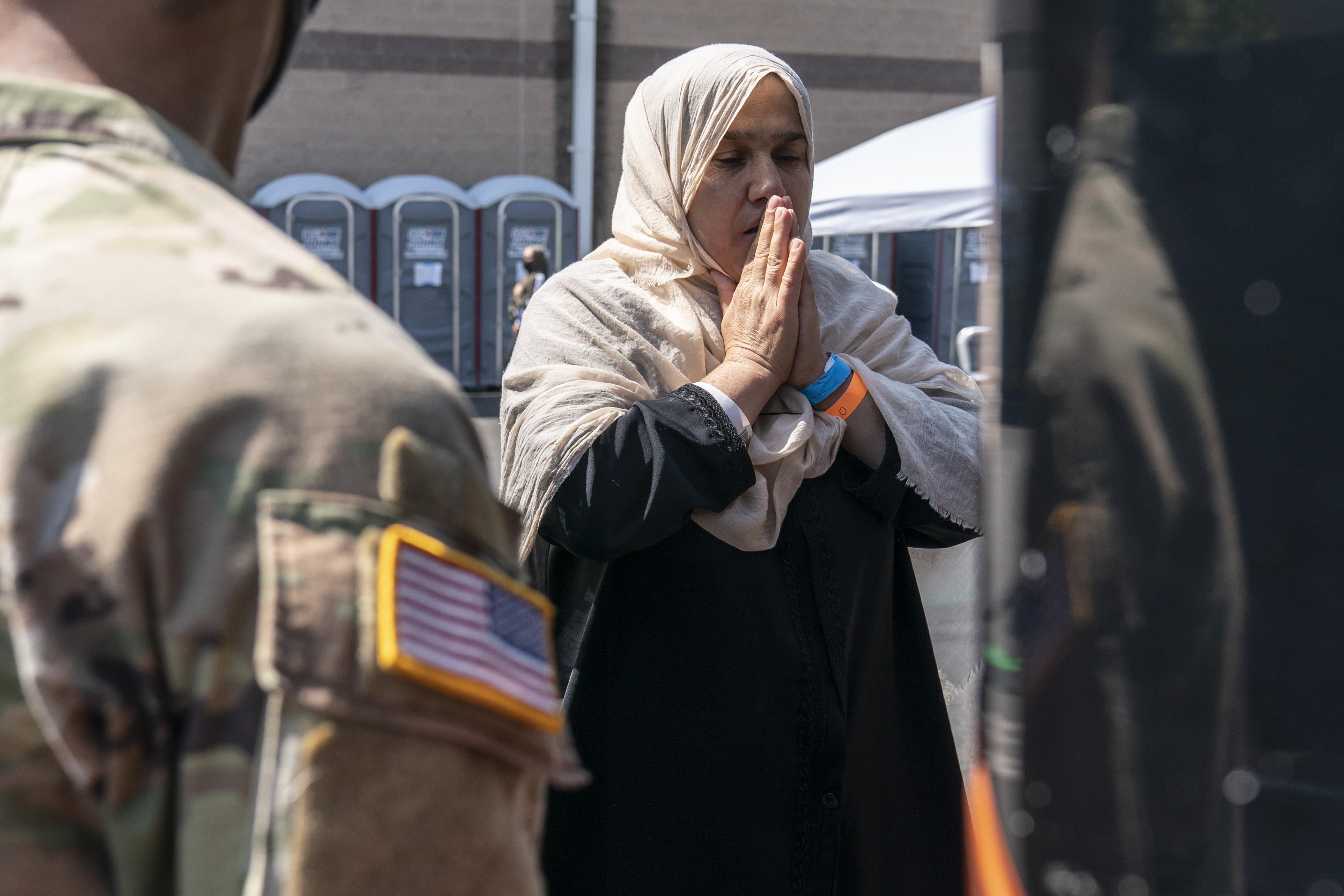 A woman stands with her hands clasped in prayer next to a bus. A U.S. service member in fatigues stands in the foreground, facing her.
