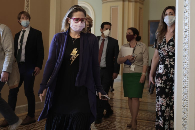 Sinema in a mask and a black dress with a lightning bolt on it walking through a hallway with other people