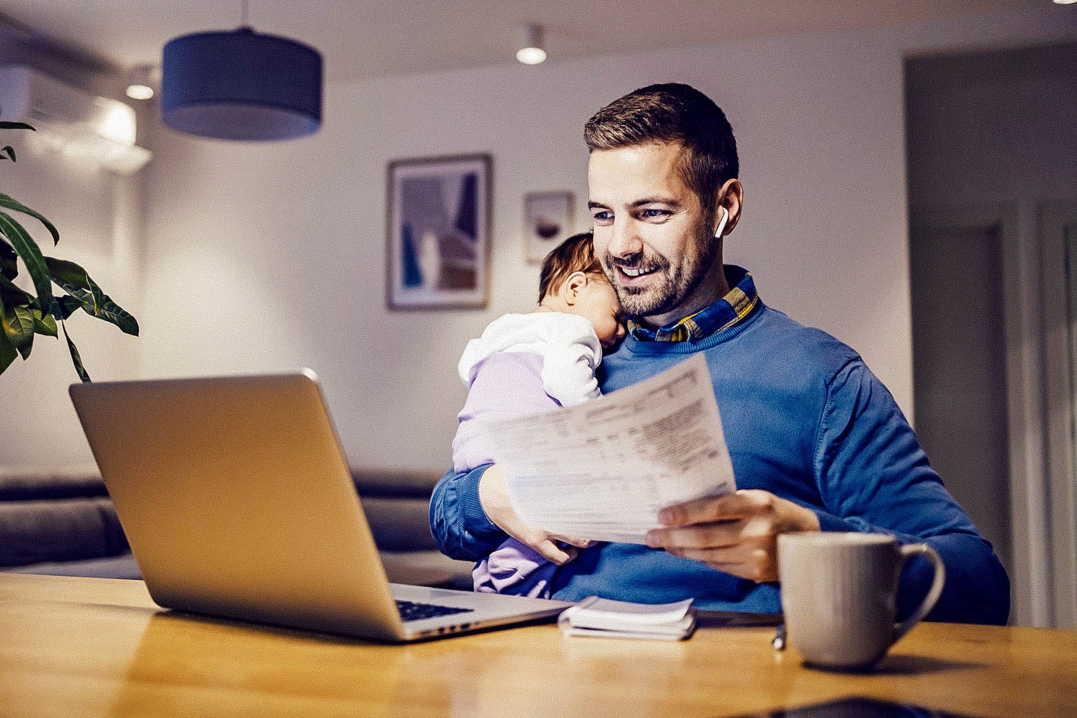 A dad smiles as he works from home sitting in front of a laptop with a baby on his shoulder and a paper in his other hand