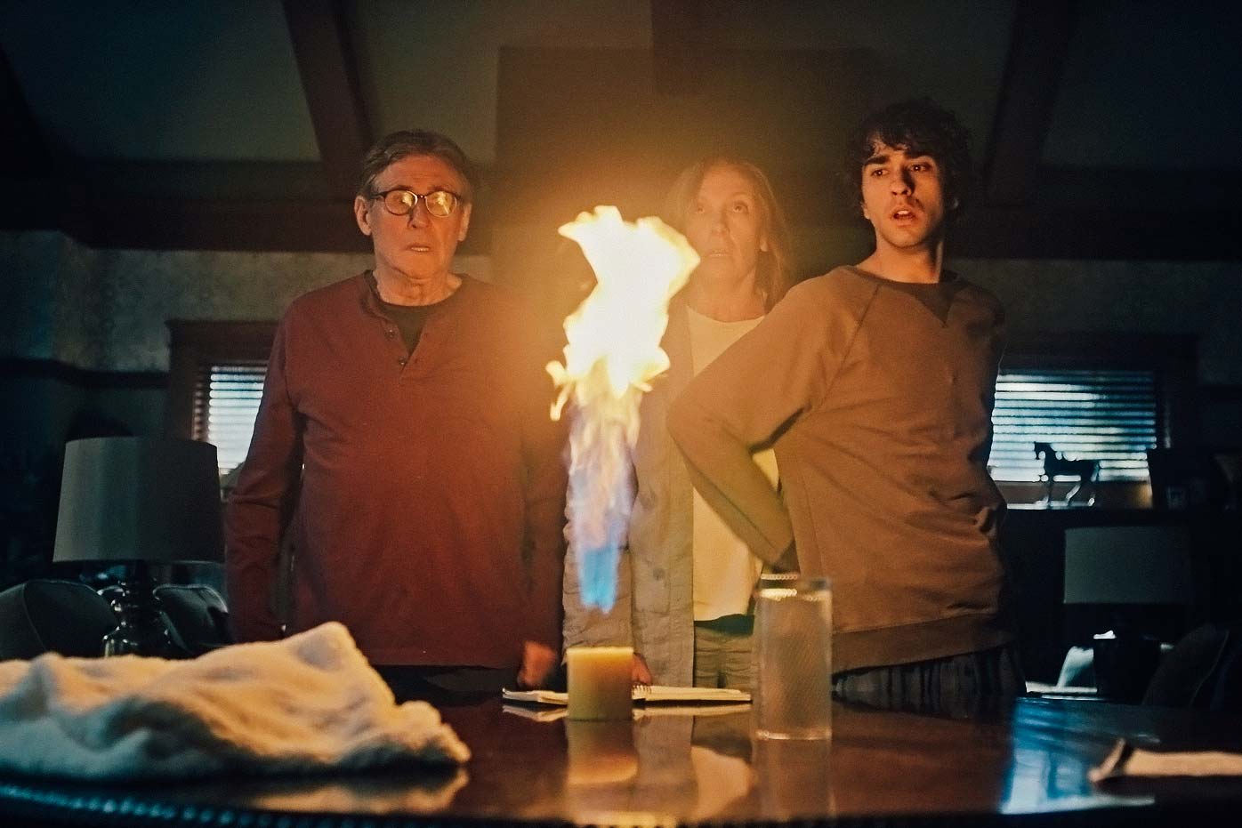 Gabriel Byrne, Toni Collette, and Alex Wolff stand around a table with an object on fire on top in Hereditary.