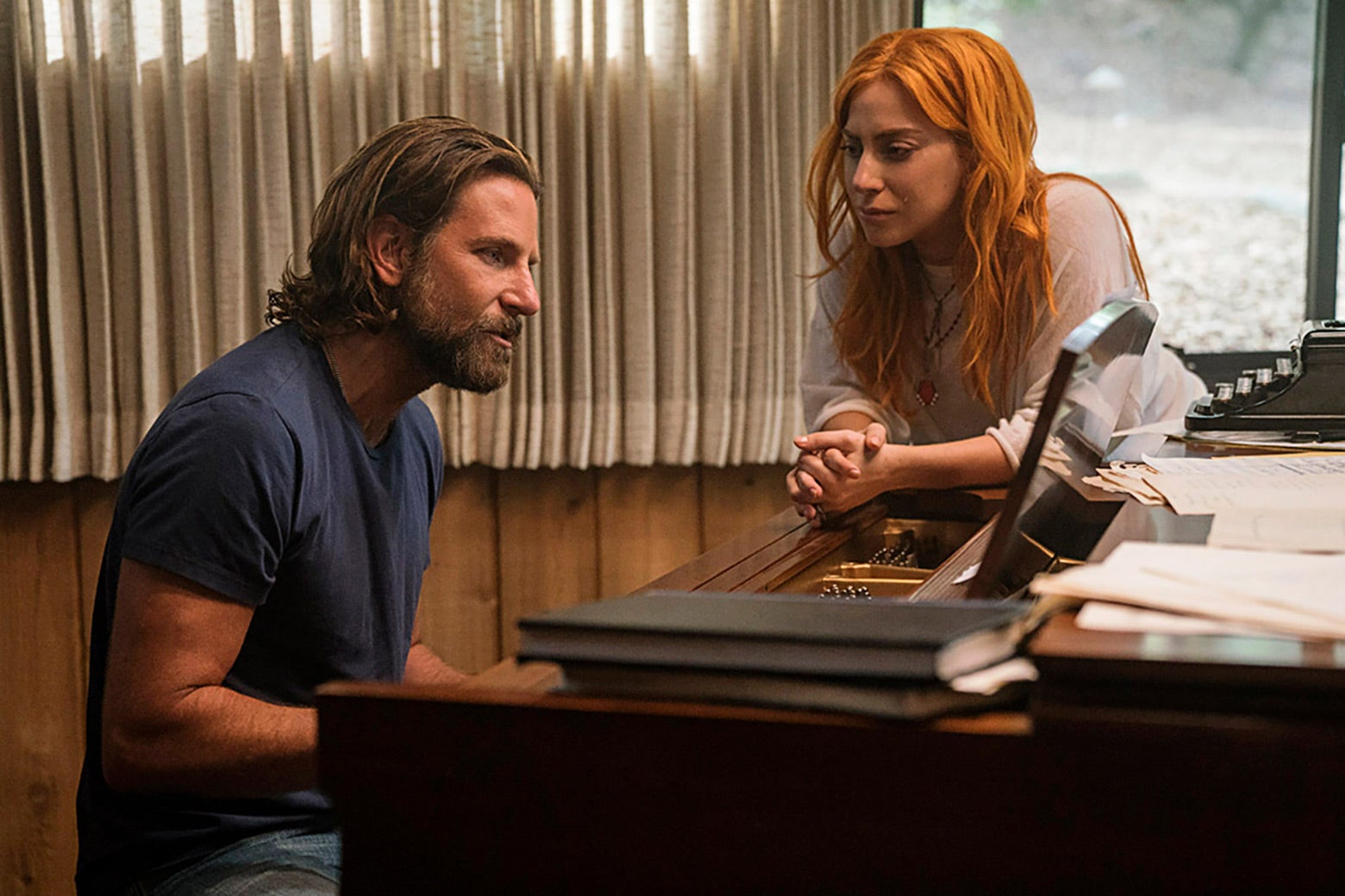 Bradley Cooper plays at the piano as Lady Gaga leans over.