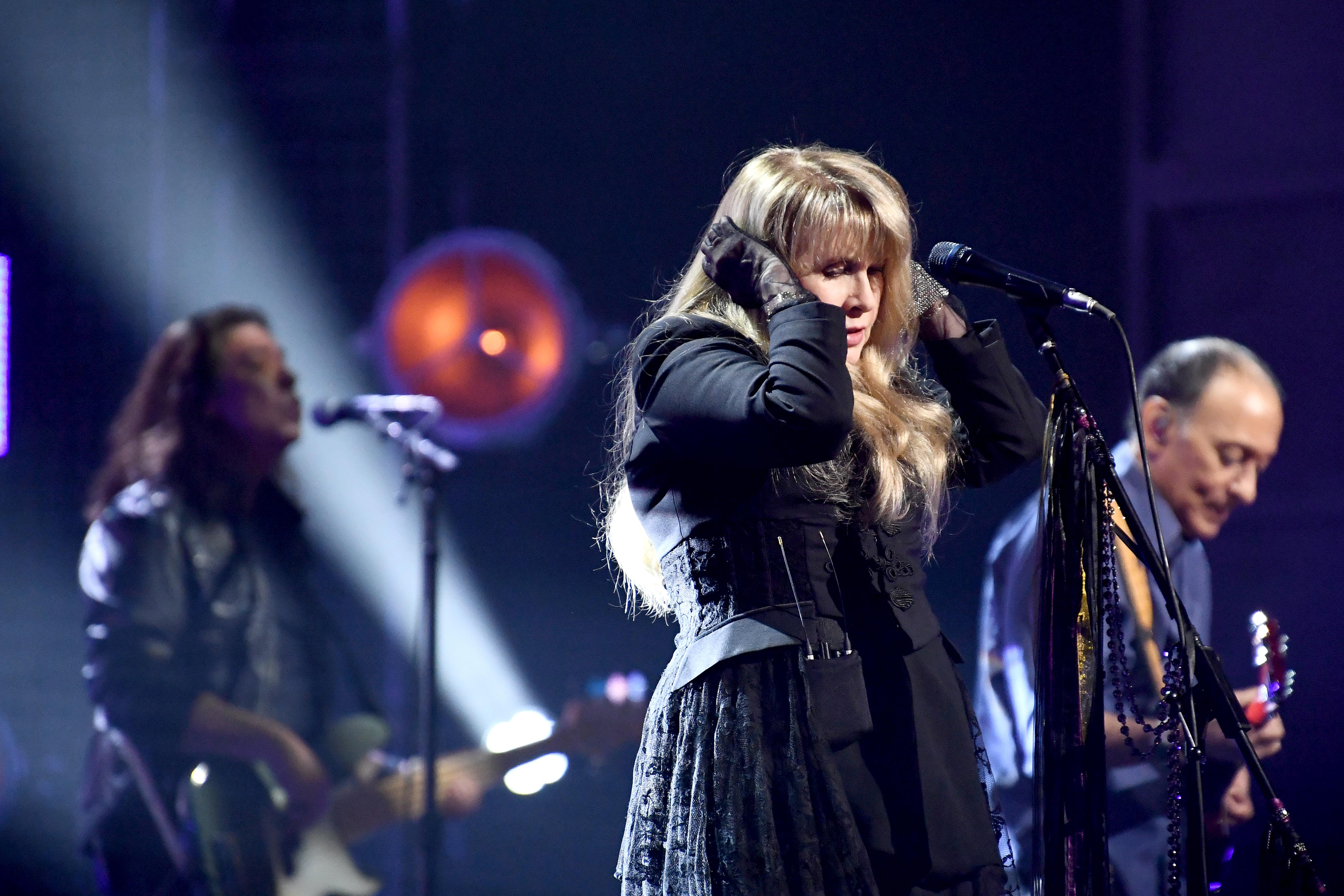 A singer puts her hand on her head while singing. Two male guitarists are shown blurry in the background of the image.