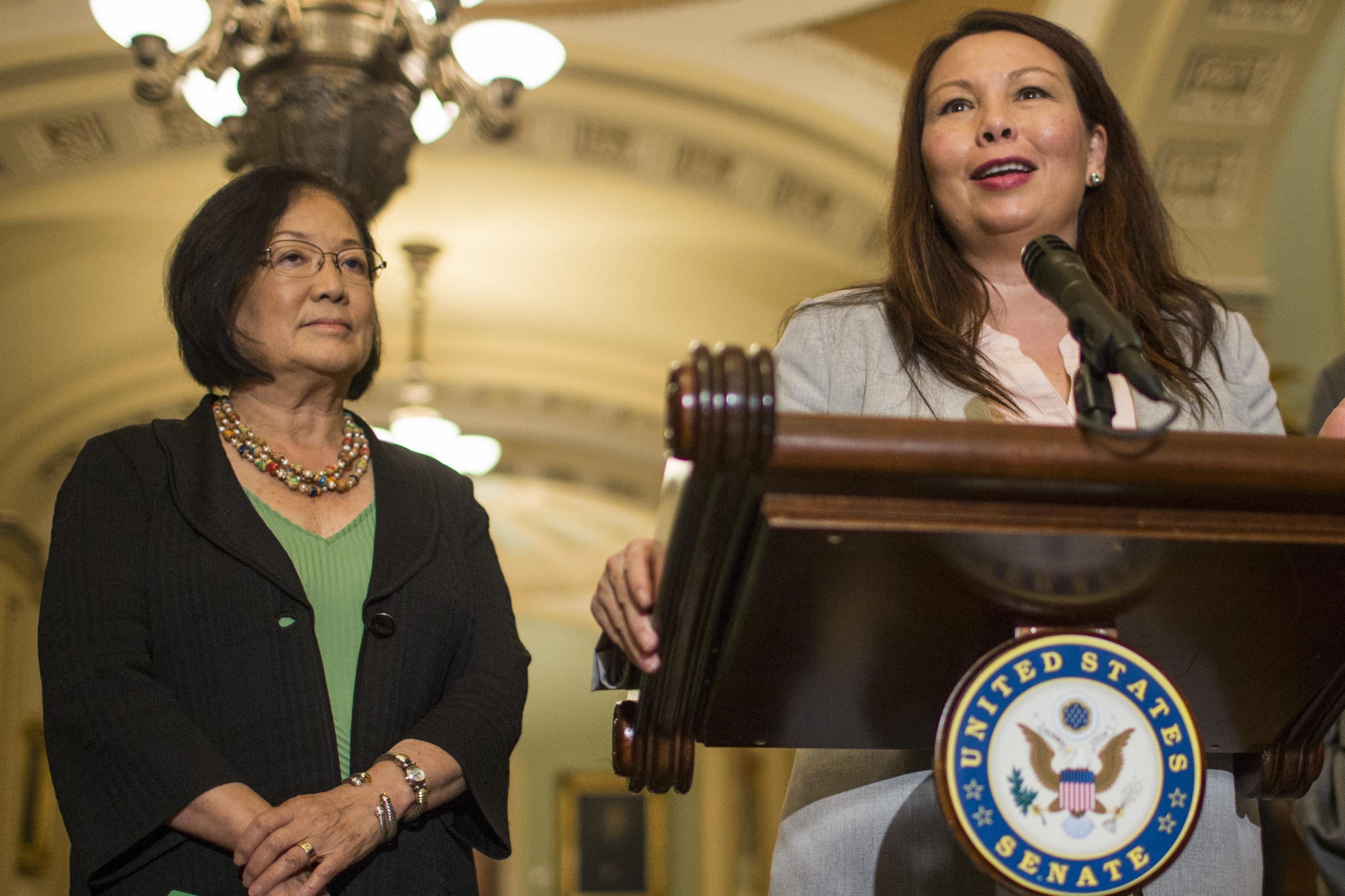 Mazie Hirono stands beside Tammy Duckworth who is speaking at a podium inside the Capitol