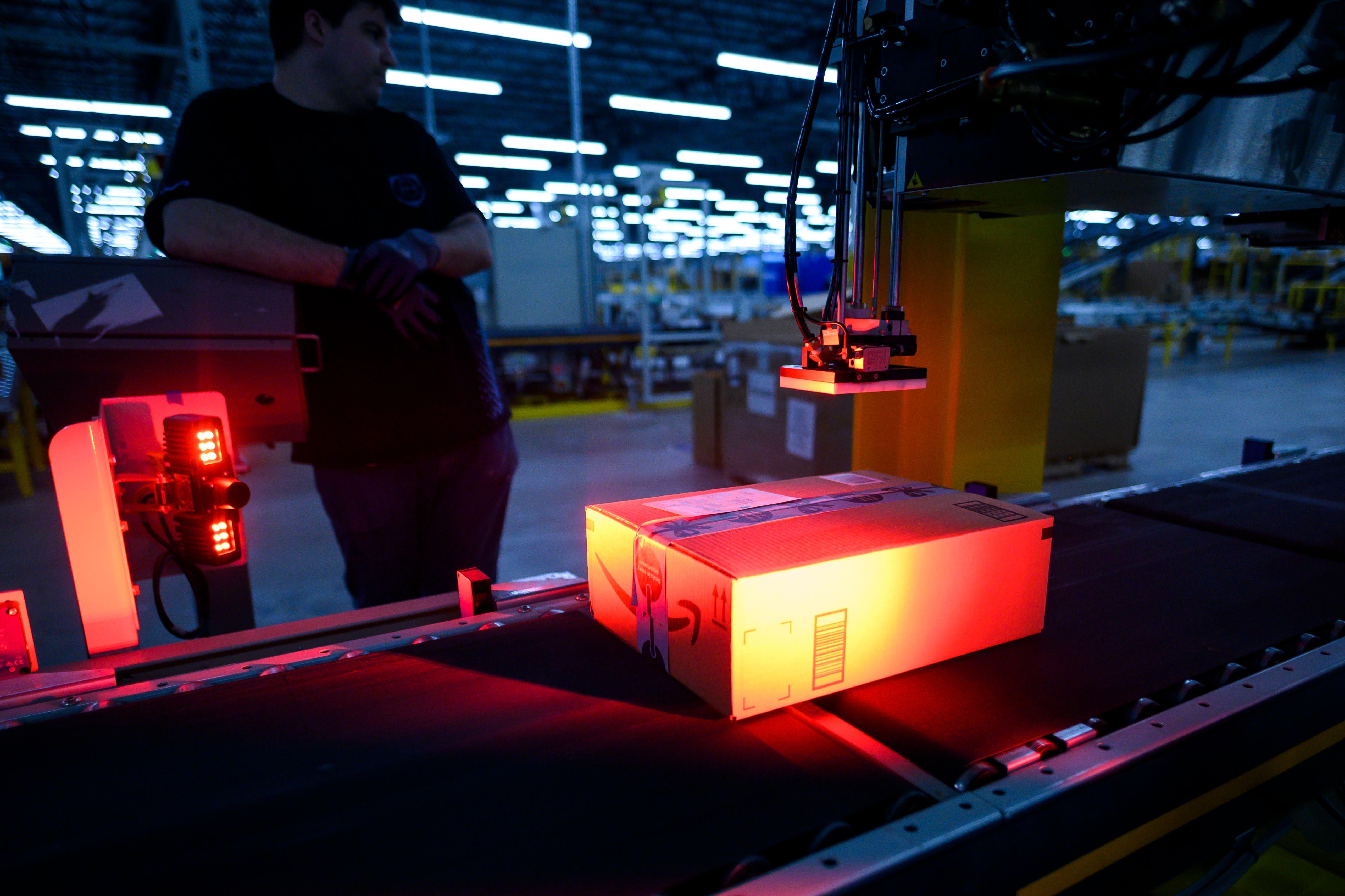 An Amazon box on a conveyor belt with a worker standing nearby in the warehouse