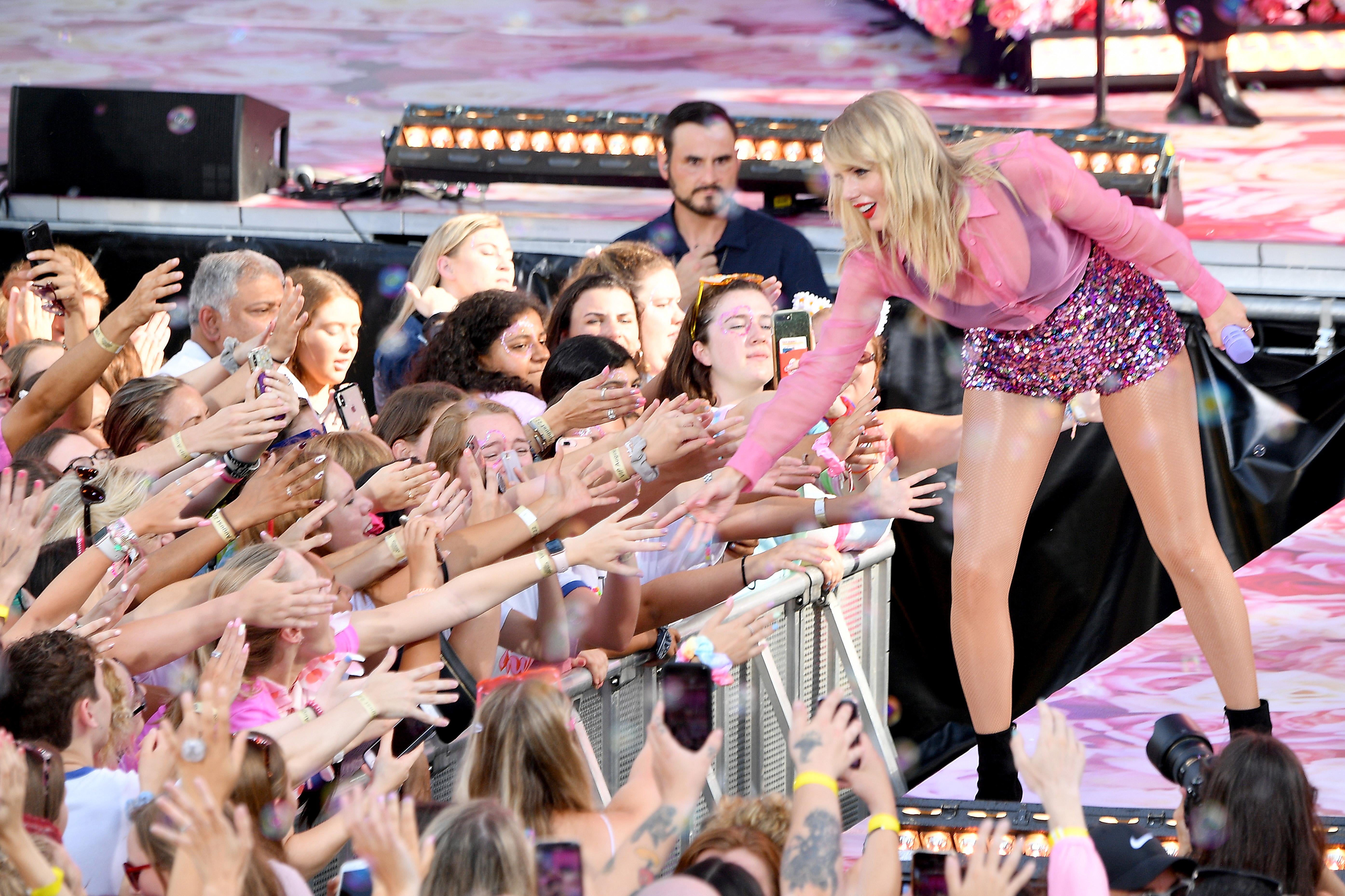 Taylor Swift leans down from a stage to touch fans' hands.