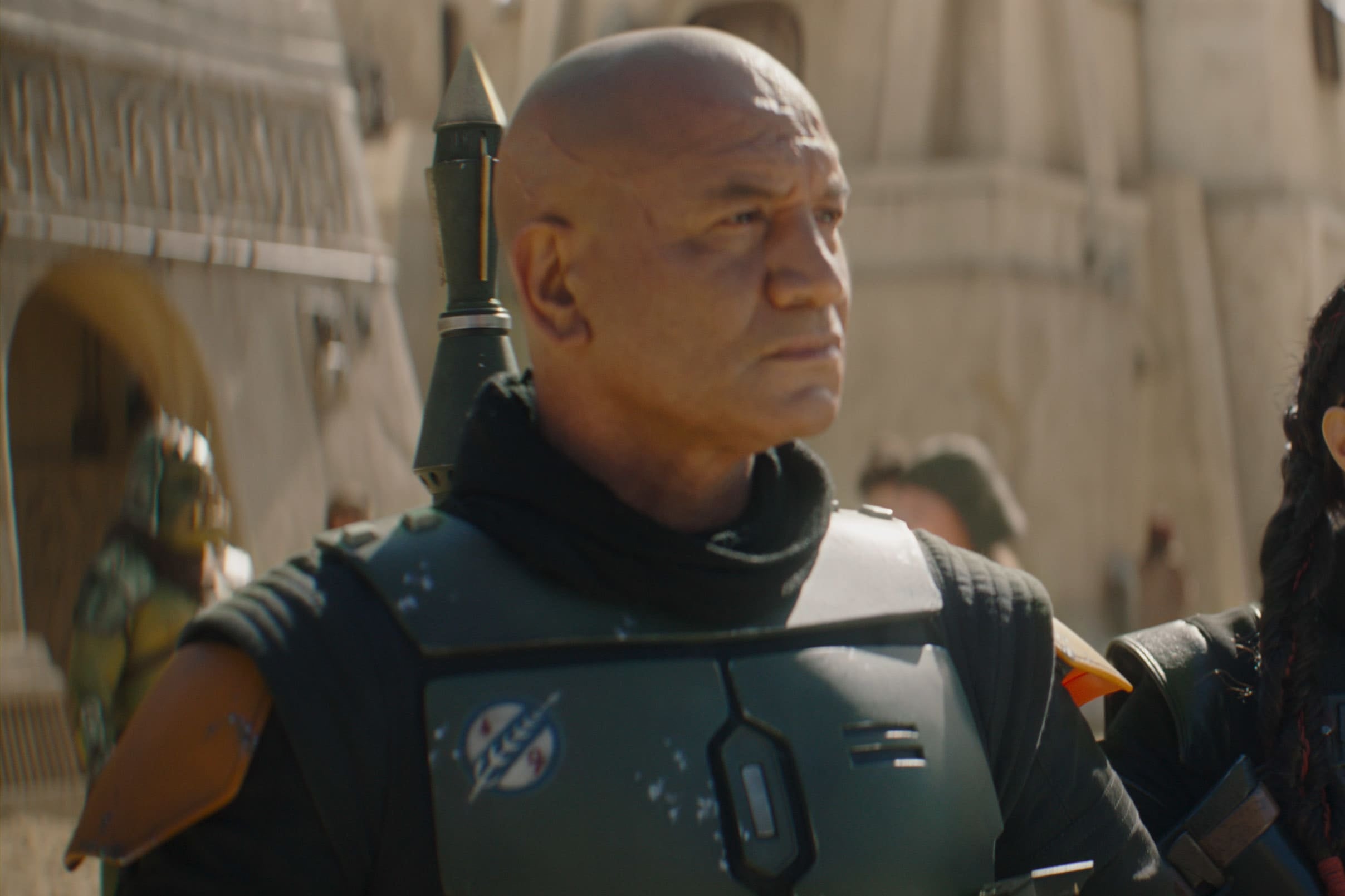 Temuera Morrison, as Boba Fett, stands in the foreground with Ming-Na Wen as Fennec Shand standing slightly behind him.  