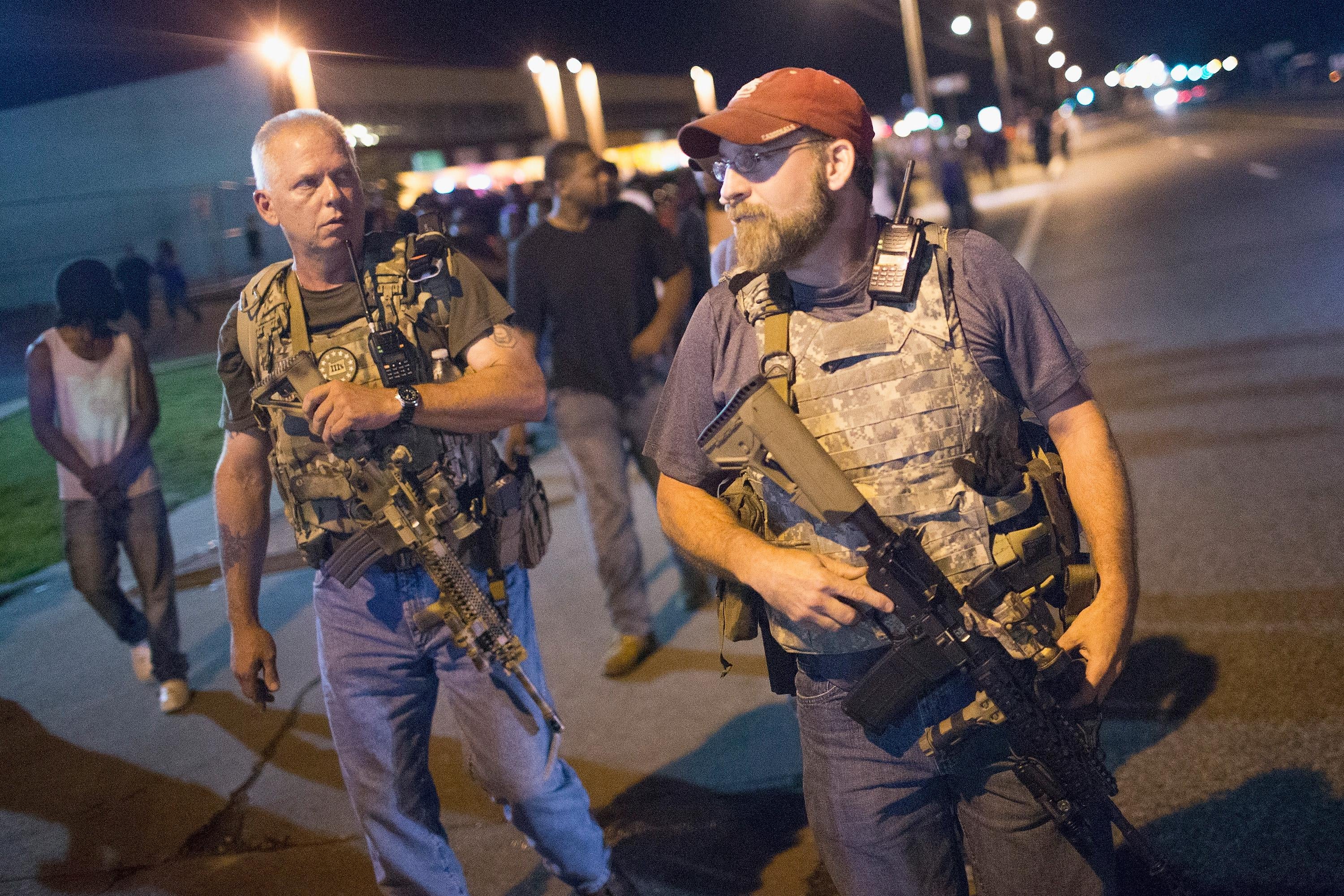 Two white men wearing camouflage vests and carrying big guns walk down a street at night