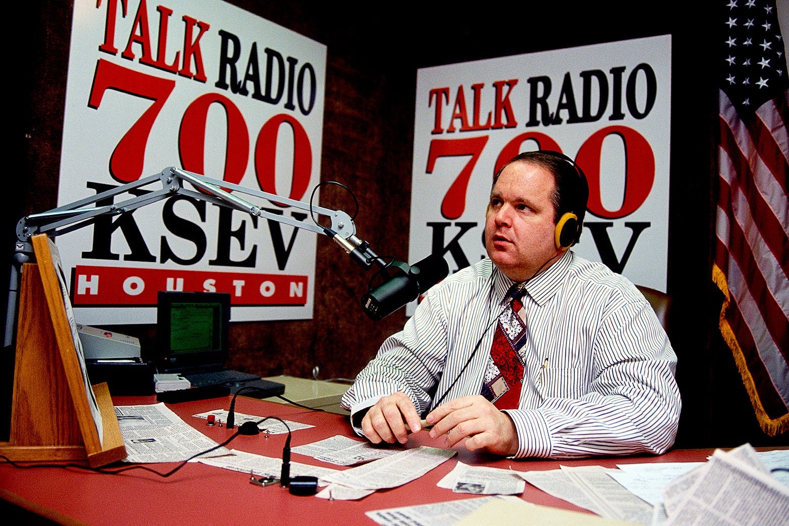 Rush Limbaugh wearing headphones, sitting in front of a mic at a desk covered in newspaper clippings