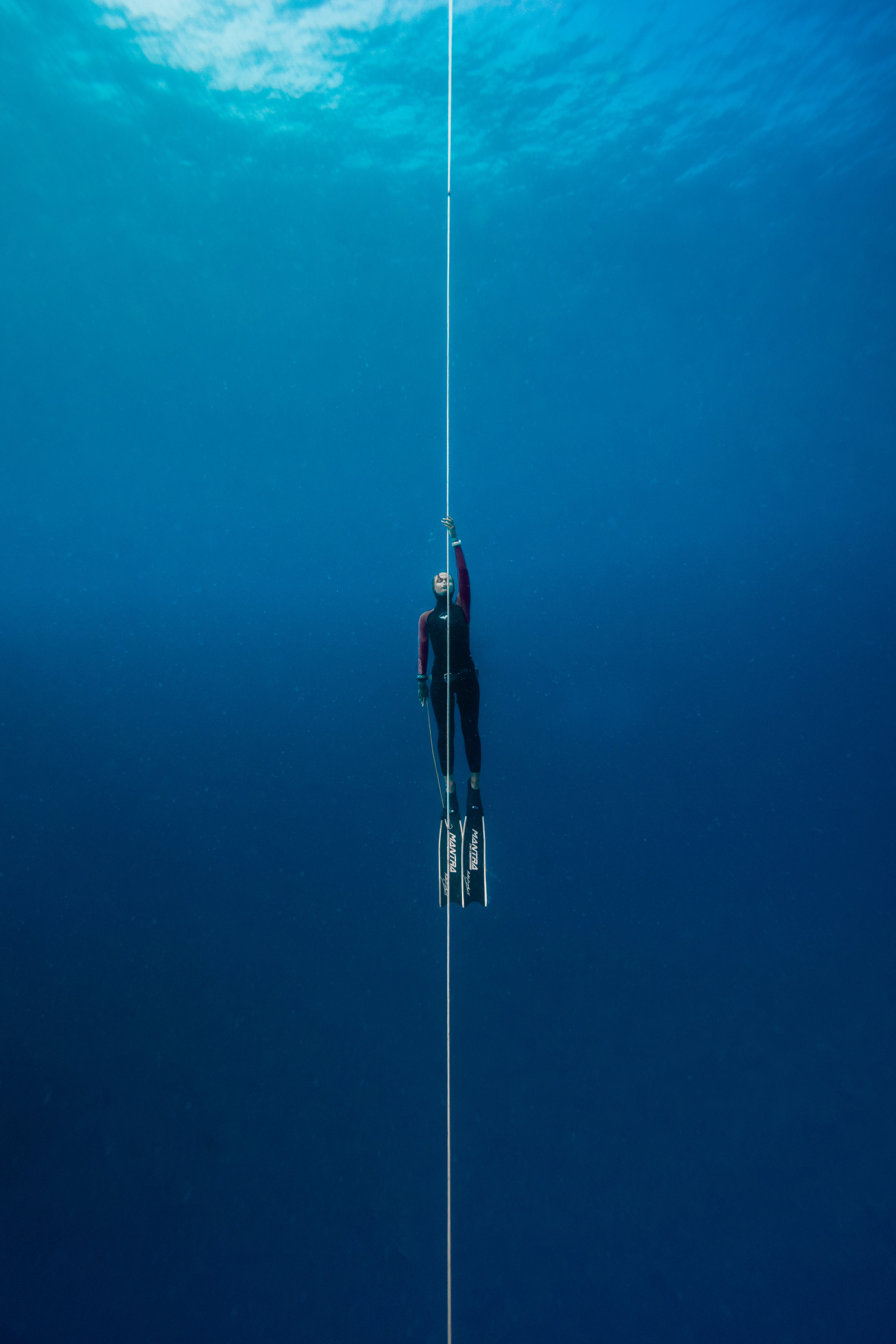 A person in a wetsuit is shown underwater pulling themselves upward via a knotted rope.