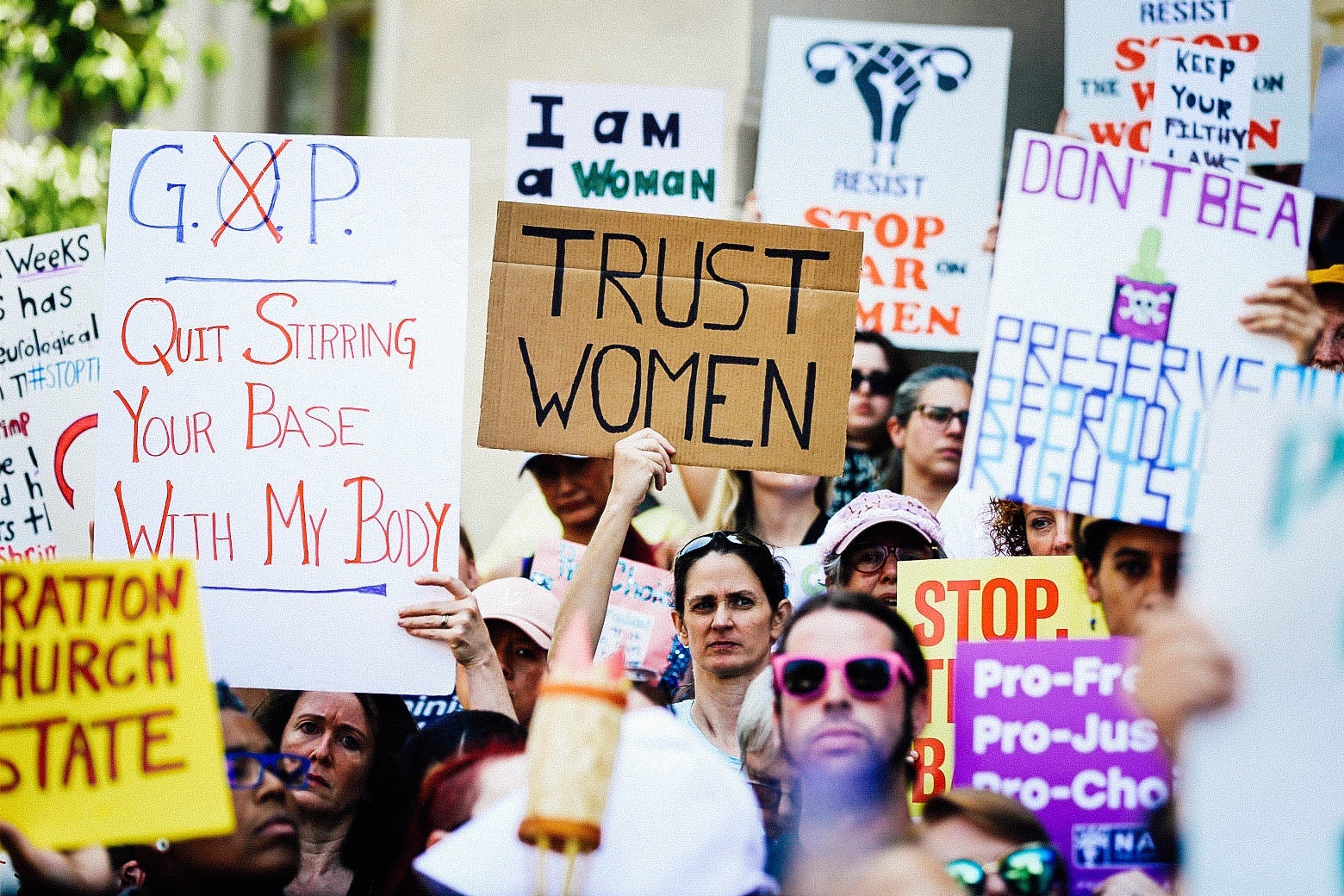 Protesters hold signs opposing the abortion ban: "TRUST WOMEN," "GOP QUIT STIRRING YOUR BASE WITH MY BODY," and more.