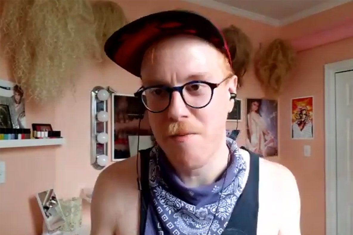 Chris Giarmo in a tank top, bandanna, glasses, and ball cap with pink walls and blond wigs in the background.