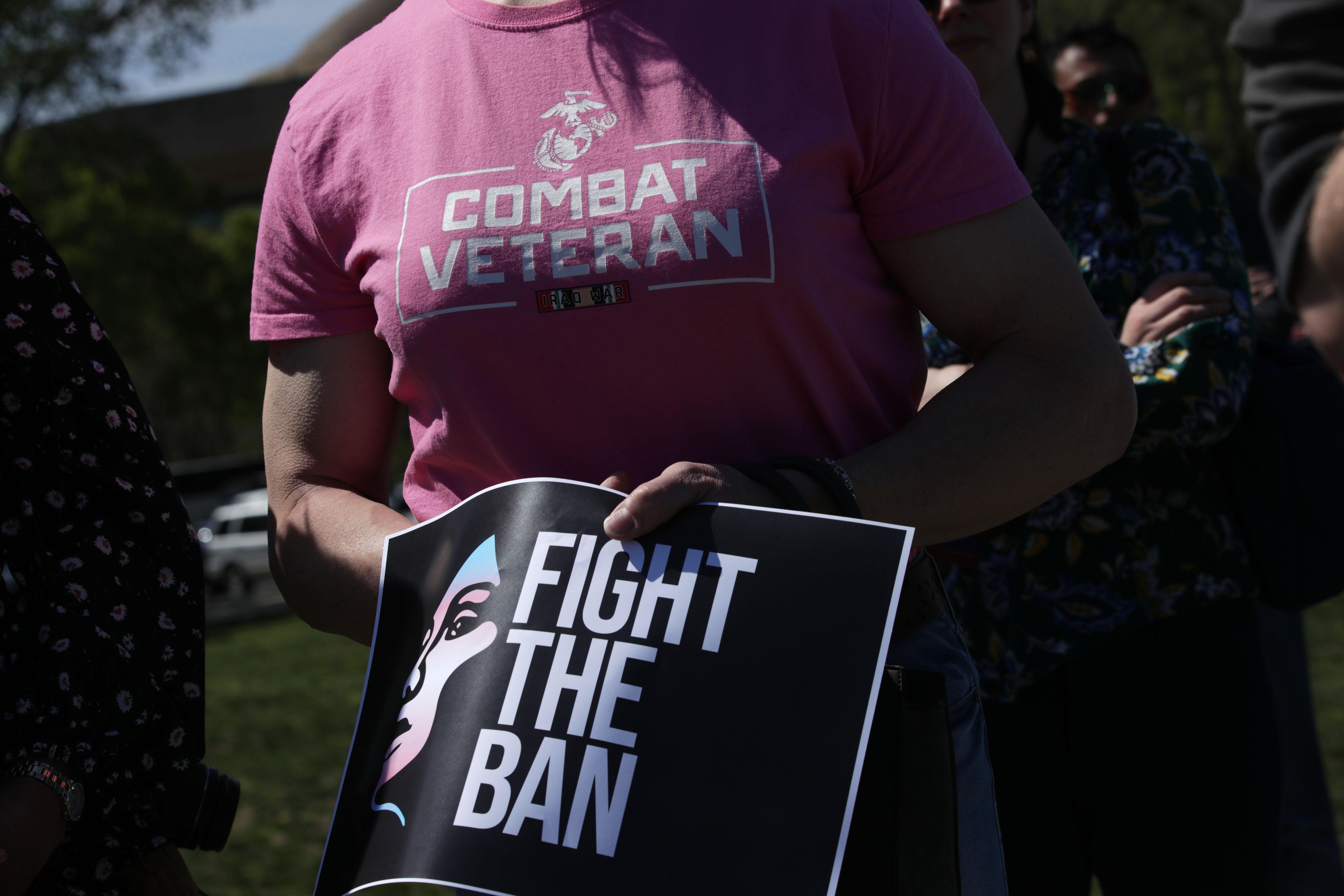 A person wearing a "Combat Veteran" T-shirt holds a sign that says "Fight the Ban."