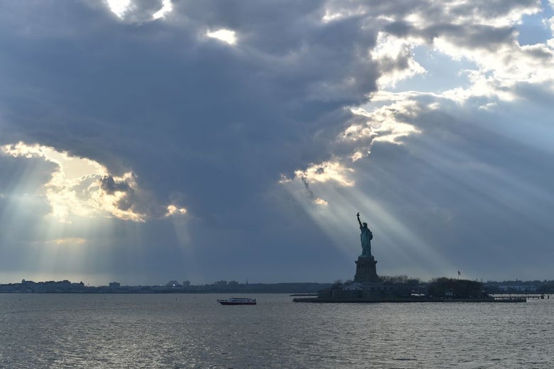 Rays of sunshine shine on the Statue of Liberty and New York Harbor from behind a cloud in the late afternoon.