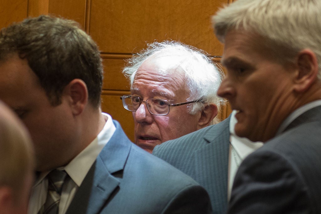 Sanders looks toward the camera with a beleaguered expression on his face in a crowded elevator.