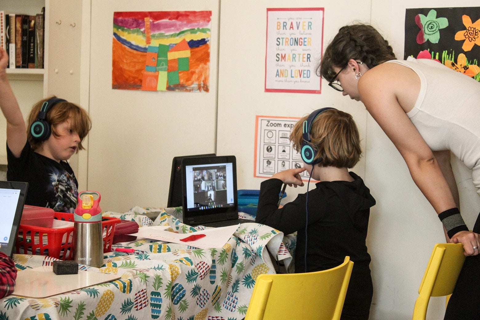 Two boys sit at laptops with headphones on as a woman hovers over.
