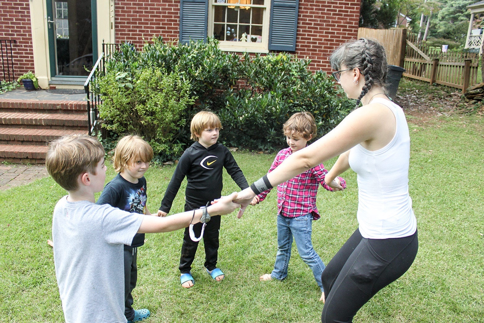 Four white children and a white woman hold hands in a circle in a lush green yard.