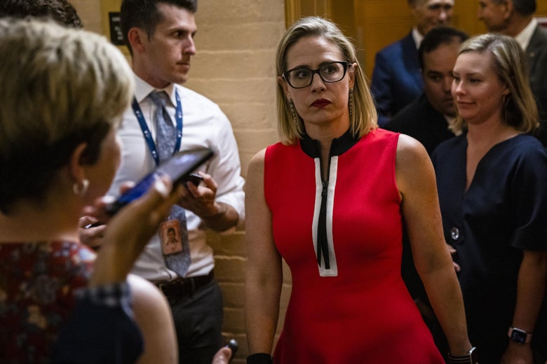Kyrsten Sinema, wearing a red dress with a large zipper, heads back into a meeting in the basement of the Capitol. 