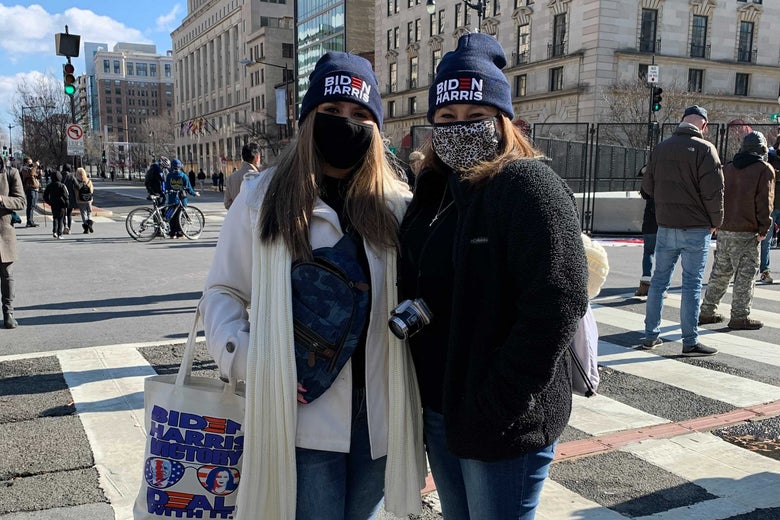 Two women wearing Biden-Harris hats stand in a crosswalk