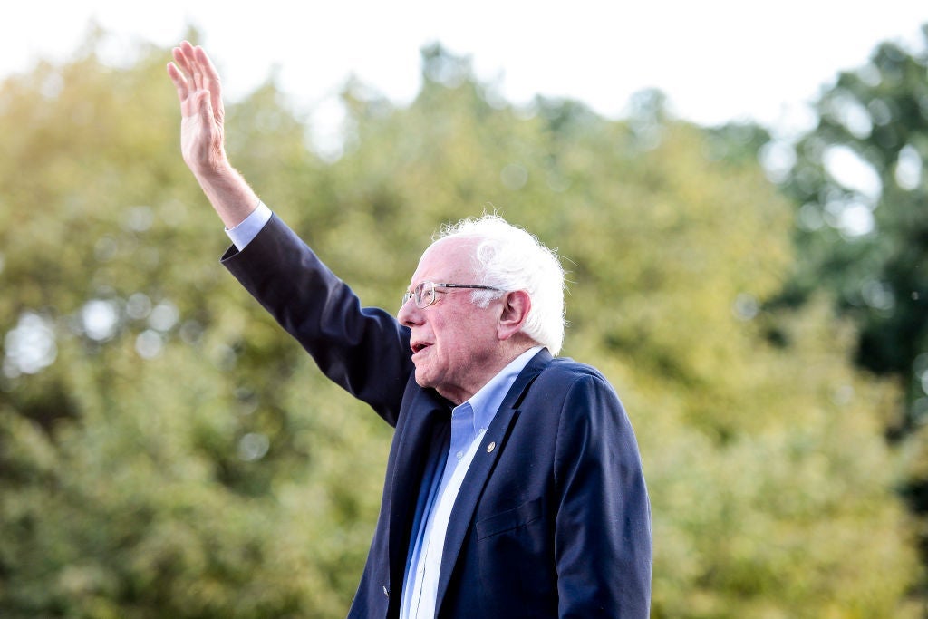 Sanders, wearing a blue suit without a tie, waves to a crowd against a backdrop of trees.