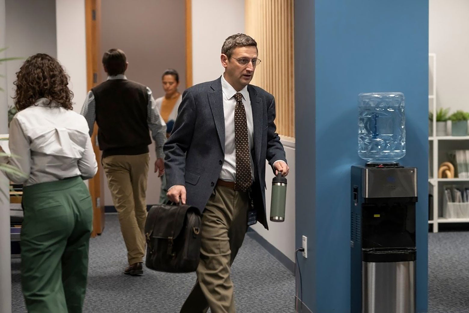 A nerdy-looking middle-aged man strides into his office, carrying a briefcase.