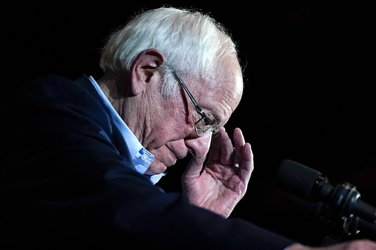 Bernie Sanders, viewed in profile, adjusts his glasses while standing behind a microphone.