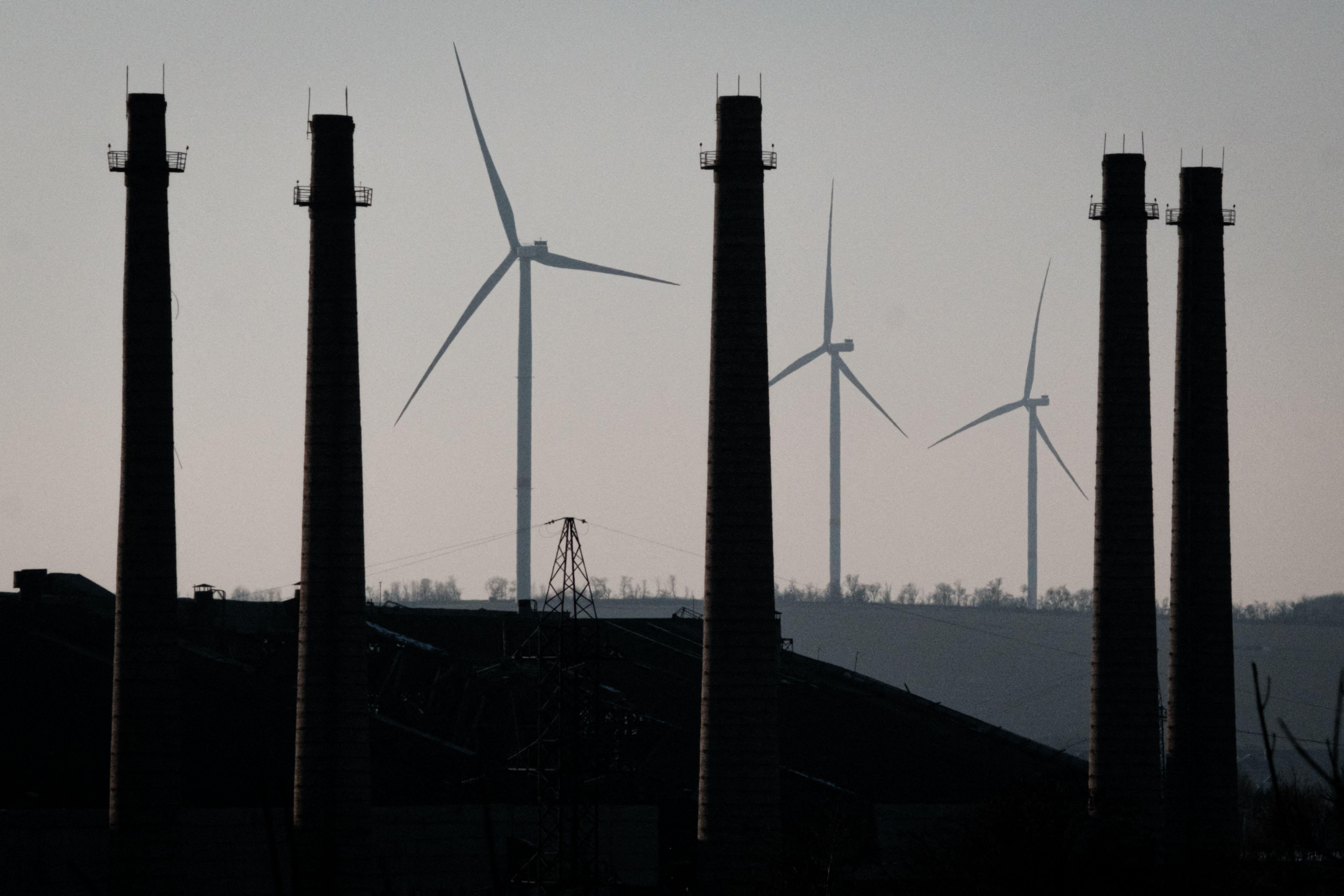 Three wind turbines with rotating blades are seen behind a group of four smokestacks. 