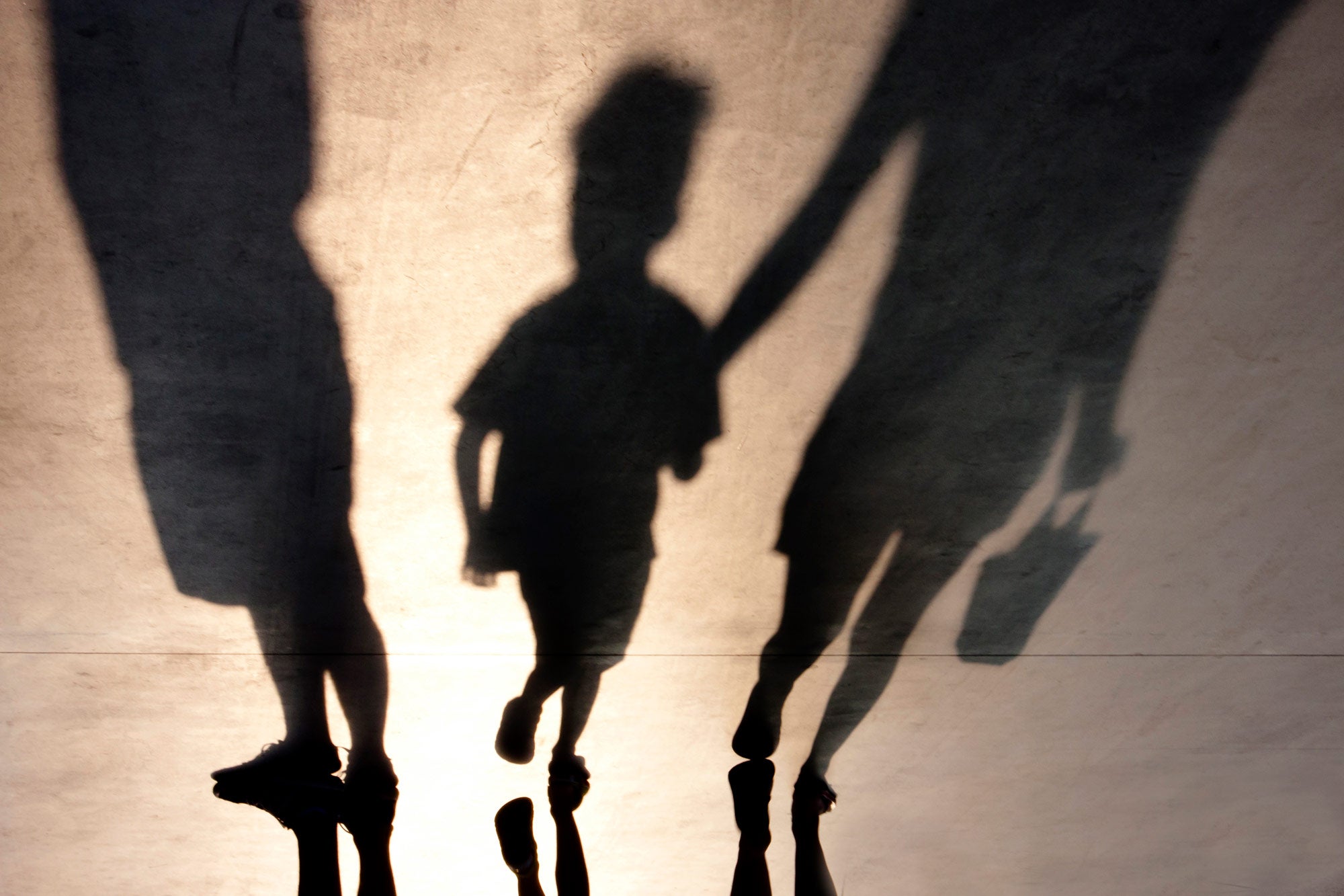 Shadows of a family being separated by parental alienation. A silhouette of a family of three with a young child or toddler holding the hand of one of the parents and being led away by them. 