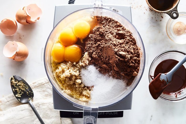 Ingredients for the cake (eggs, cocoa, quinoa, sugar) sit in a food processor next to a bowl of ganache.