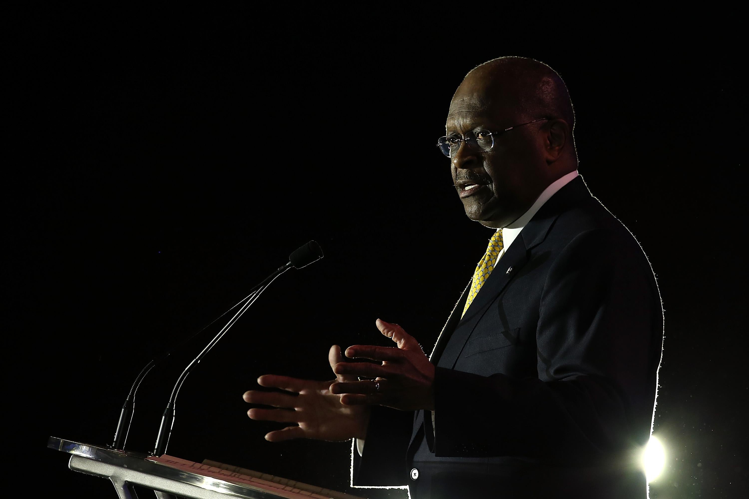 Cain speaks at a podium on a dark stage