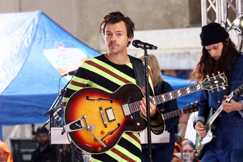 Harry Styles performs with a Gibson guitar on the Today Show, looking straight at the camera, wearing swoopy hair and a skintight bodysuit that looks like a very 1970s candy cane