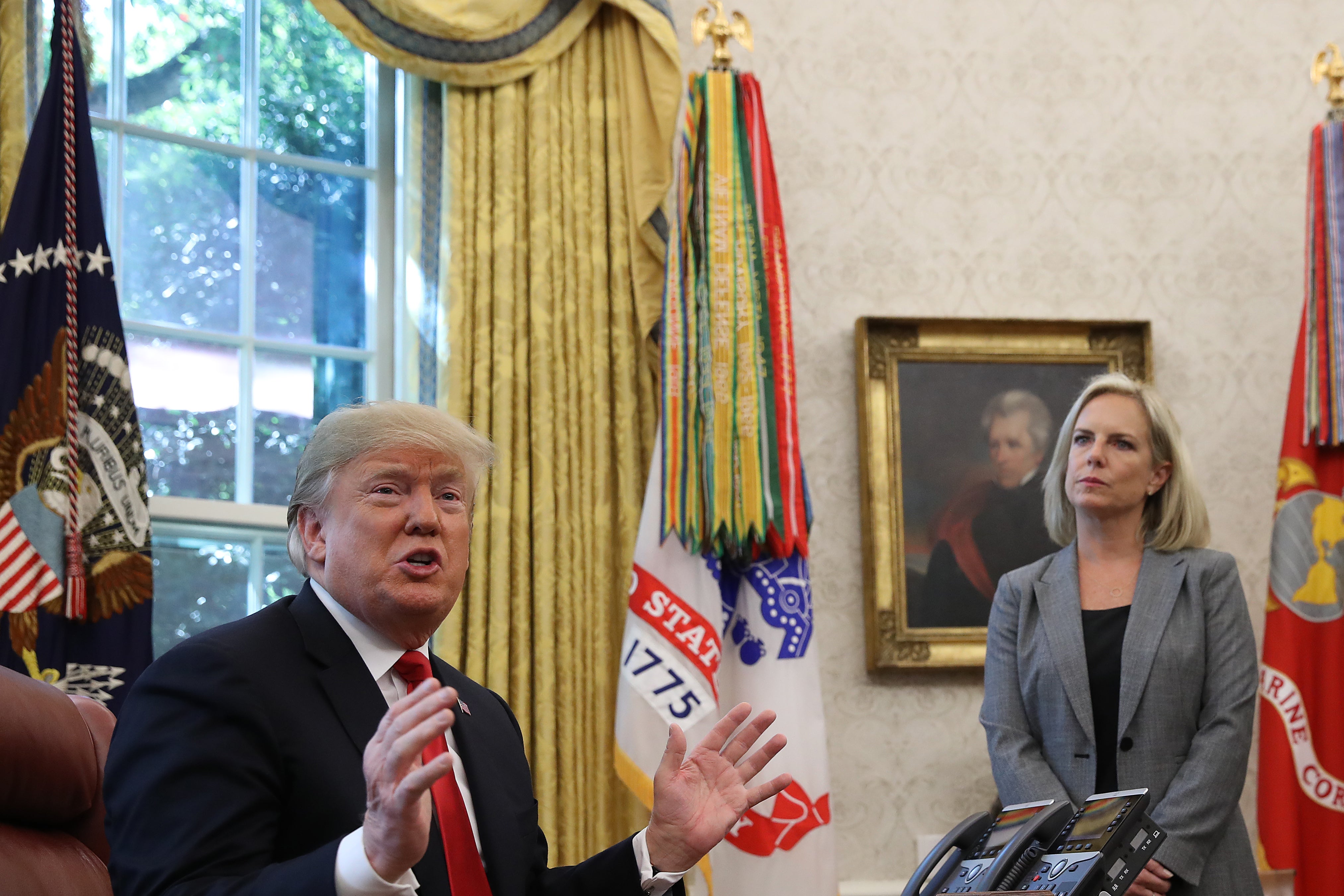 Trump, seated at a desk, gestures while speaking. Nielsen stands in the background.