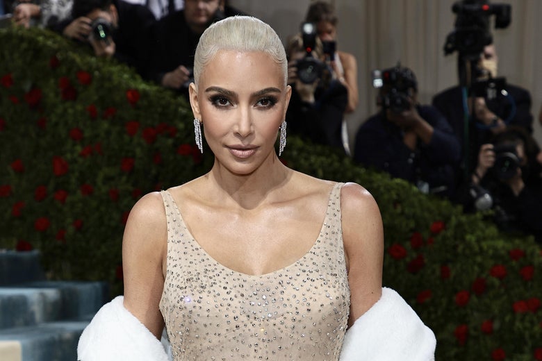 Kim Kardashian in a beige dress, with slicked-back blonde hair. She is on a red carpet, posing with a neutral-but-seductive expression. A row of photographers are behind her.